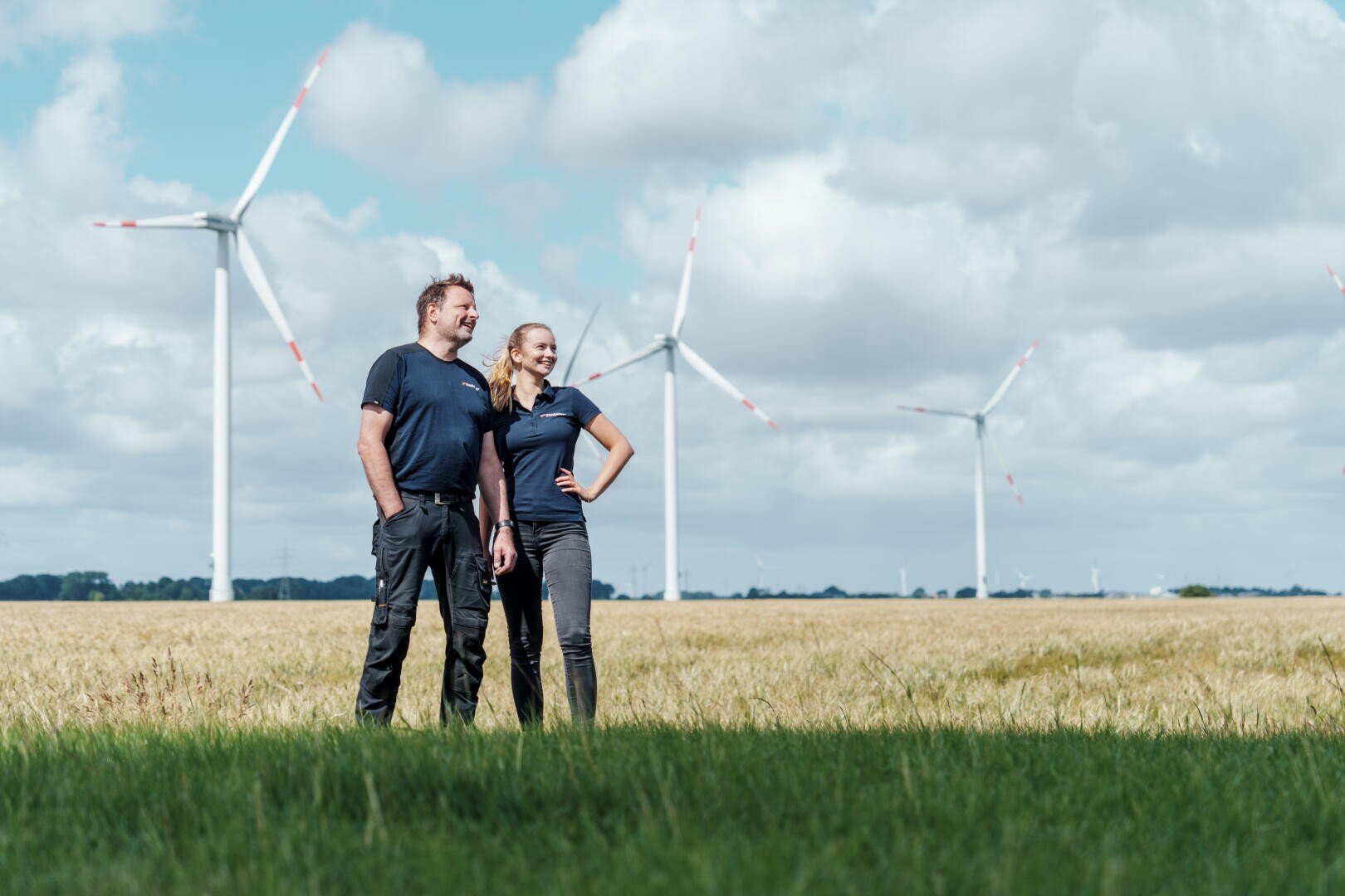 Ein Mann und eine Frau stehen zusammen auf einer Wiese mit Windrädern im Hintergrund und einem wolkenverhangenen blauen Himmel über ihnen, lächeln und schauen in die Ferne.