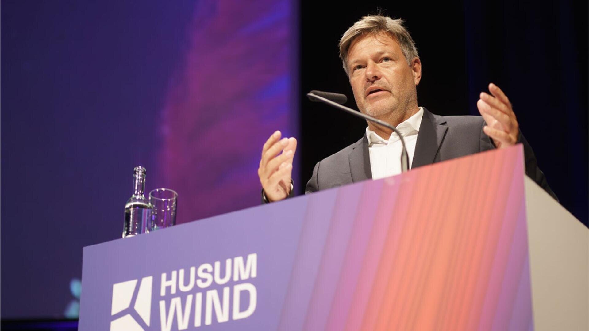 A man in a suit speaks at a podium labeled Husum Wind, gesturing with his hands. A microphone, glass, and bottle are visible, with a purple backdrop behind him.
