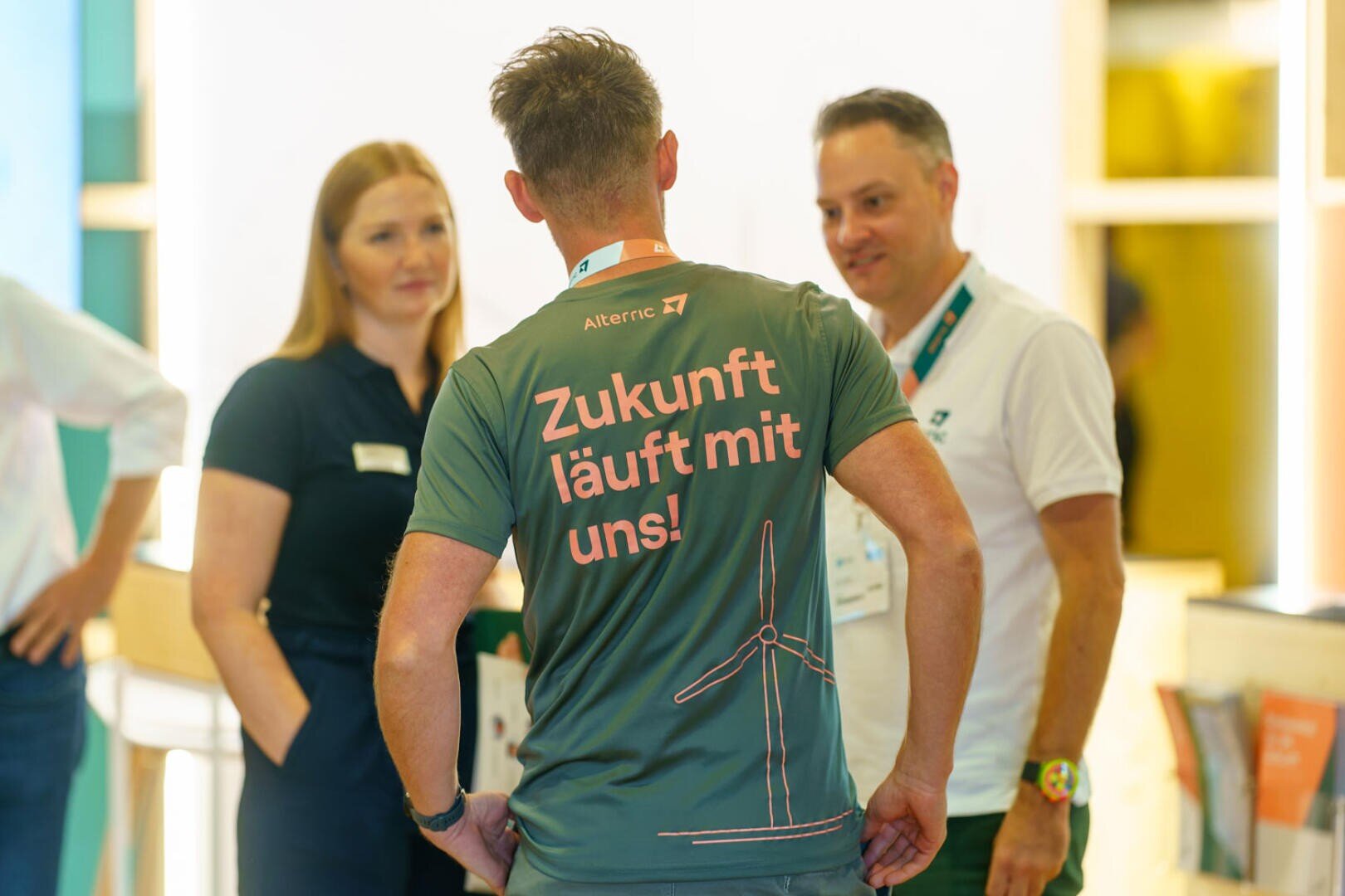 Ein Mann, der ein Hemd mit der Aufschrift "Zukunft läuft mit uns!" und einer Windturbinengrafik trägt, unterhält sich bei einer Indoor-Veranstaltung mit zwei anderen Personen. Die Atmosphäre wirkt freundlich und professionell.
