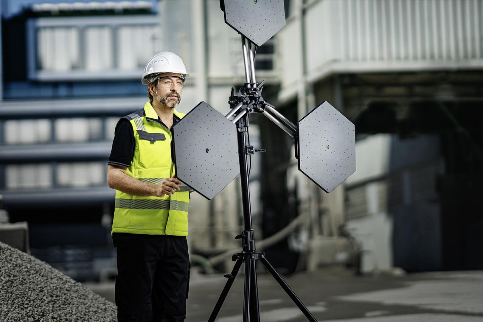 A man wearing a hard hat and neon-colored safety vest is standing outside next to a tripod with three hexagonal panels, possibly carrying out a technical or structural inspection on an industrial site.