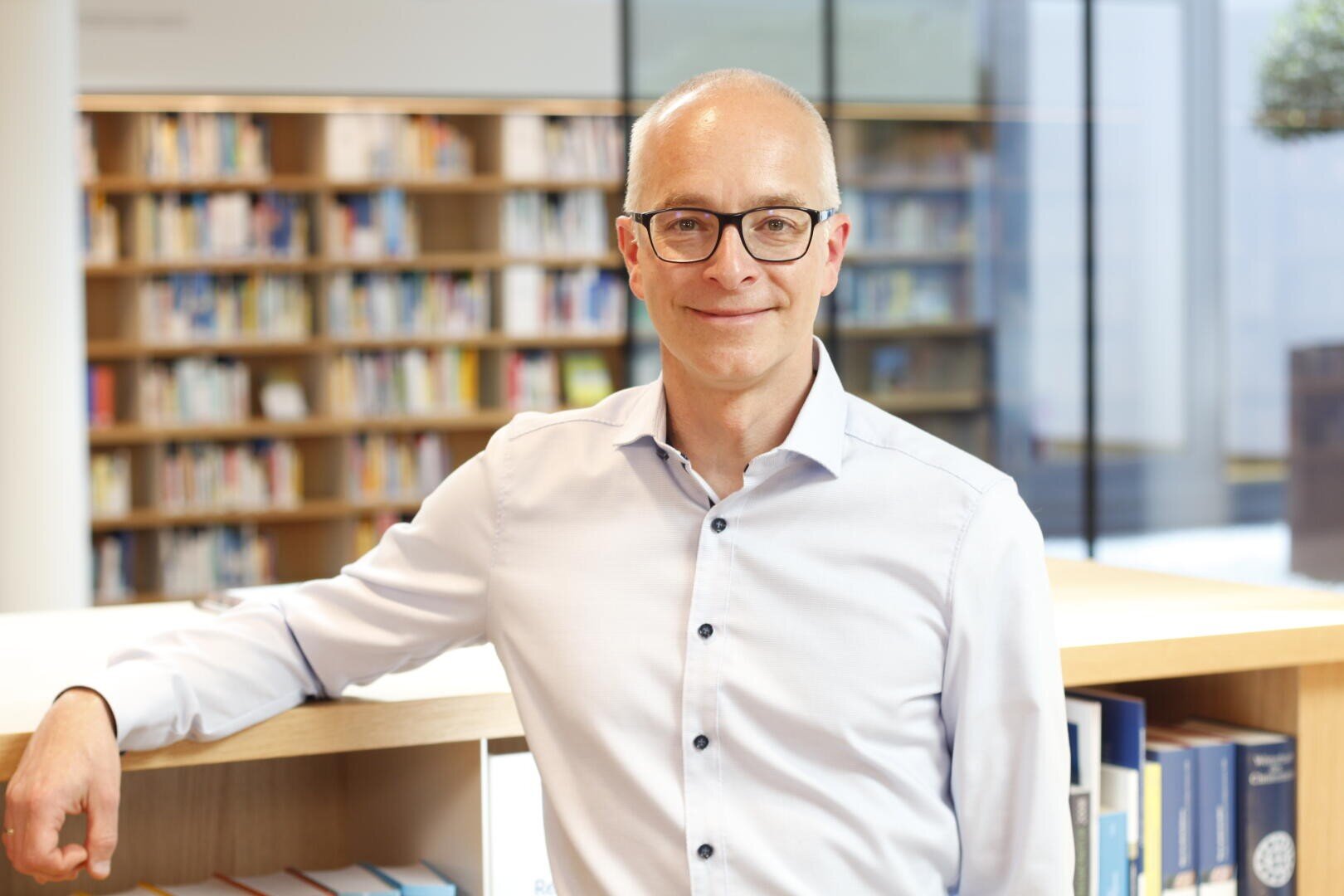 A smiling man with glasses and a white shirt stands in a modern library and rests his arm on a wooden shelf filled with books. Blurred bookshelves and large windows can be seen in the background.