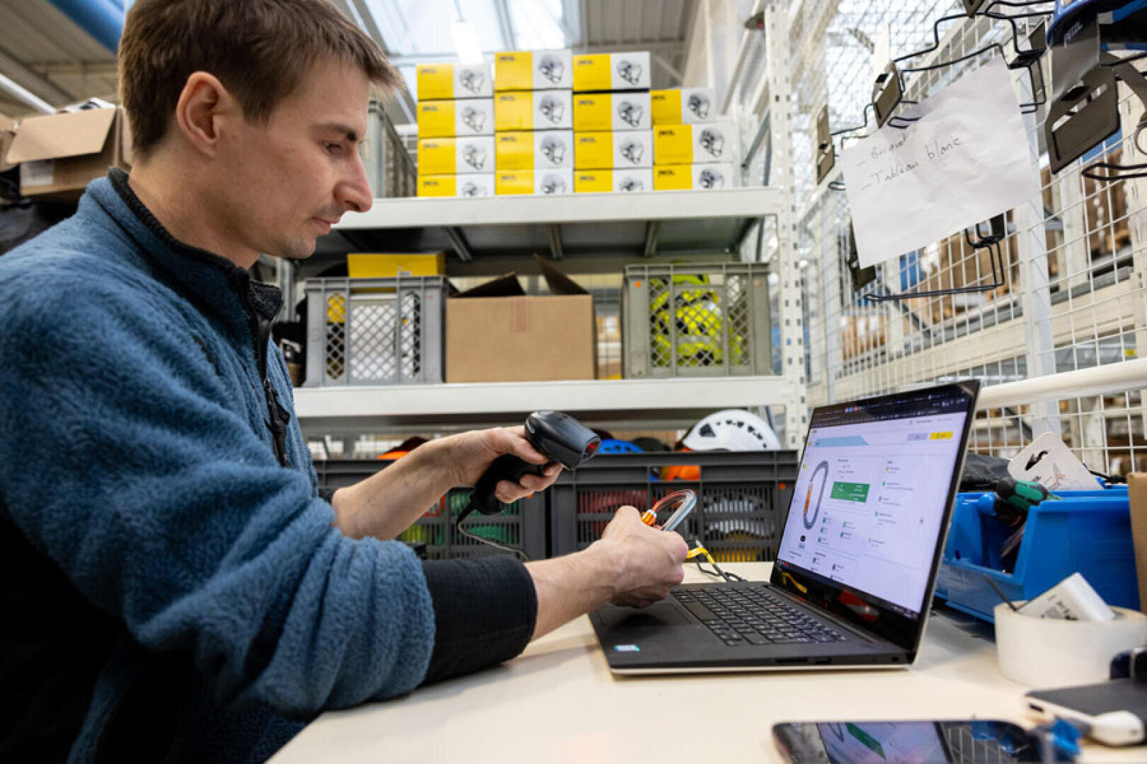 Ein Mann scannt ein Produkt mit einem Barcode-Scanner, während er an einem Schreibtisch mit einem offenen Laptop in einem Lager oder einer Werkstatt sitzt. Im Hintergrund sind Regale mit Kisten und Geräten zu sehen.