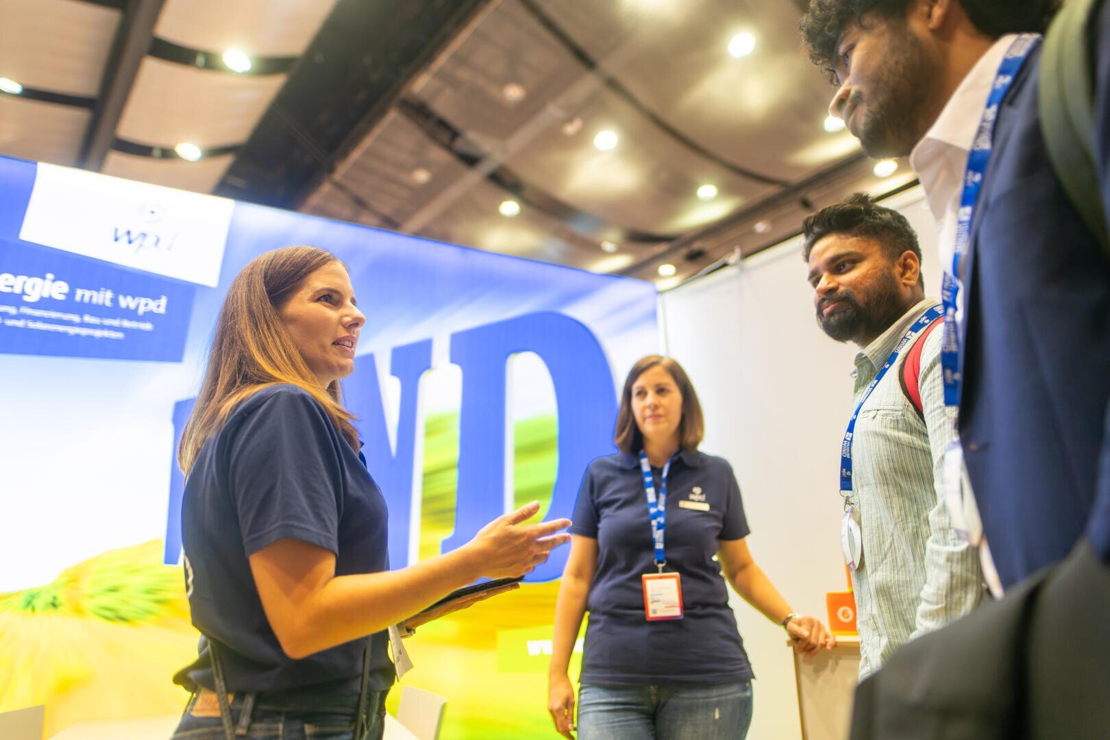Two women and two men are chatting at a stand with a blue and yellow background with the word WIND on it. They are all wearing lanyards and the environment looks professional and friendly.