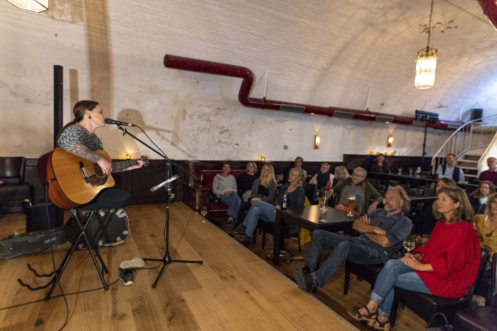 Eine Frau spielt Akustikgitarre und singt in ein Mikrofon auf einer kleinen Bühne in einem gemütlichen, schwach beleuchteten Veranstaltungsort. Mehrere Personen sitzen an Tischen und hören aufmerksam zu. Getränke stehen auf den Tischen, daneben steht ein Gitarrenkoffer.