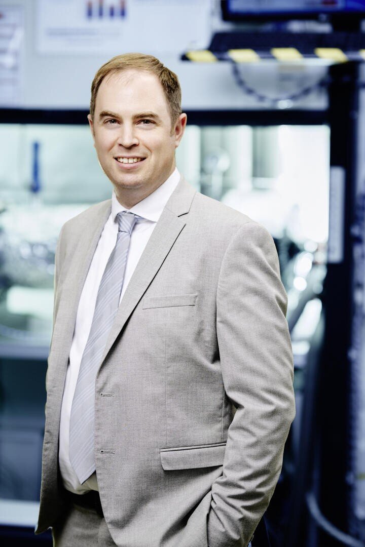 A man wearing a light grey suit and tie stands indoors, smiling at the camera. The background is slightly blurred, showing equipment and monitors, suggesting a professional or industrial setting.