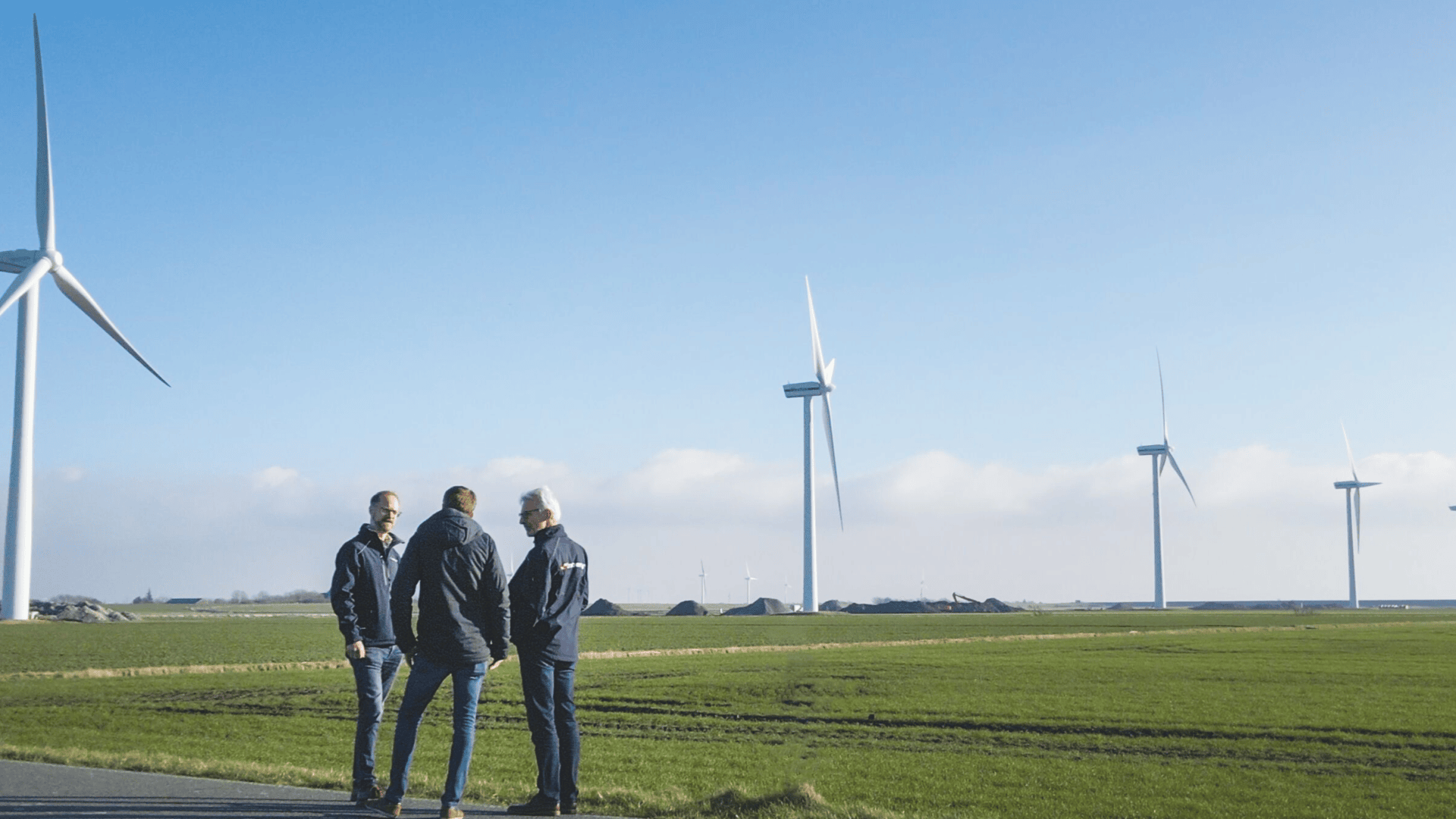 Drei Personen stehen auf einer Wiese und unterhalten sich, während im Hintergrund mehrere große Windturbinen unter einem strahlend blauen Himmel zu sehen sind.