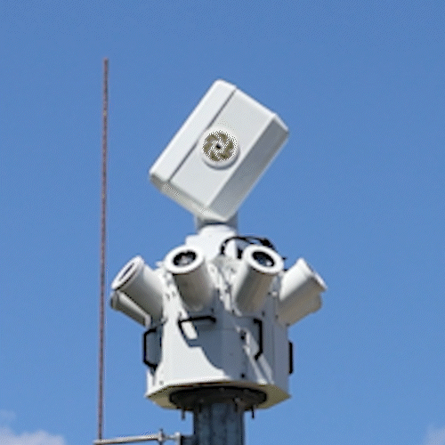Ein weißes, mehrlinsiges Überwachungs- oder Sensorgerät ist an einem Mast vor einem klaren blauen Himmel angebracht. Das Gerät hat mehrere zylindrische Sensoren und eine rechteckige Komponente an der Spitze.