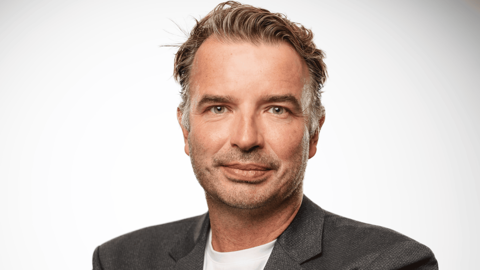 Ein Mann mittleren Alters mit kurzen, leicht zerzausten braunen Haaren, leichten Bartstoppeln und einem neutralen Gesichtsausdruck trägt einen dunklen Blazer über einem weißen T-Shirt und posiert vor einem schlichten hellen Hintergrund.