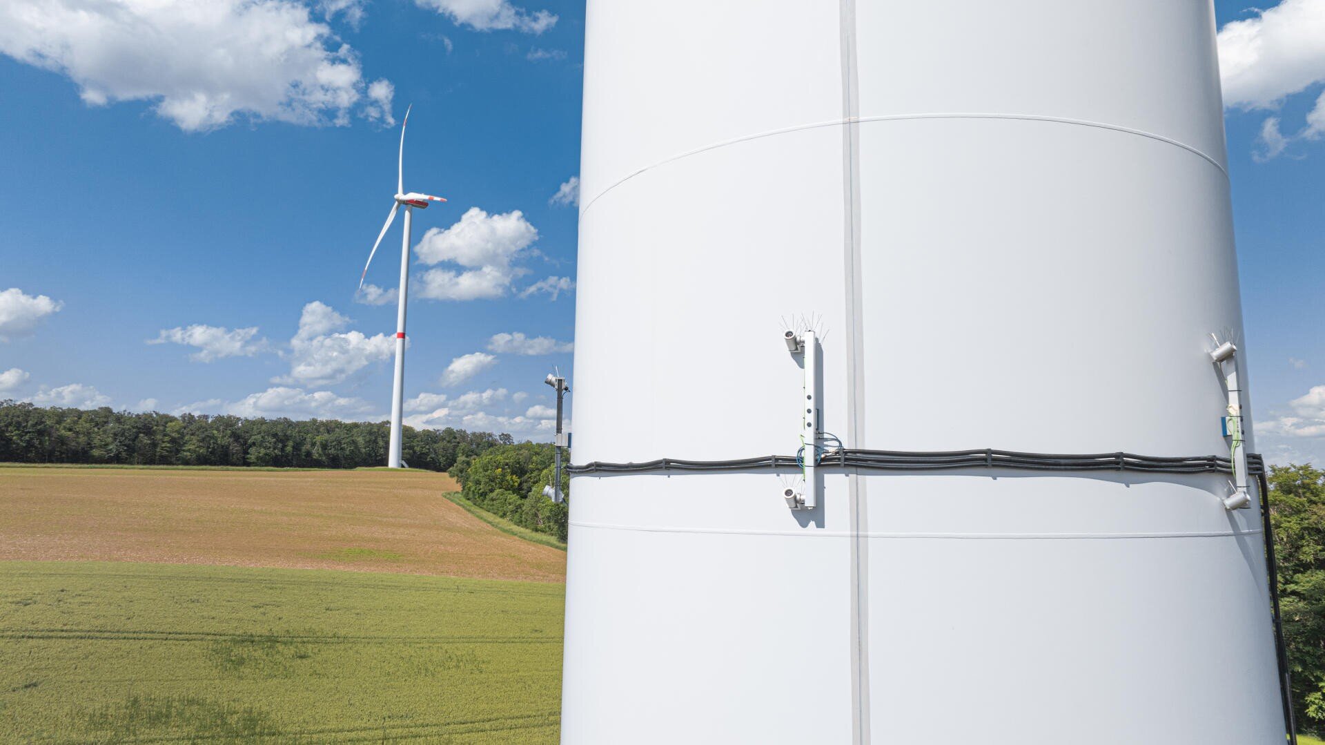 Nahaufnahme eines weißen Windradturms mit befestigten Kabeln. Im Hintergrund ist eine weitere Windkraftanlage in einem Feld mit grünen und braunen Pflanzen unter einem blauen Himmel mit vereinzelten Wolken zu sehen.
