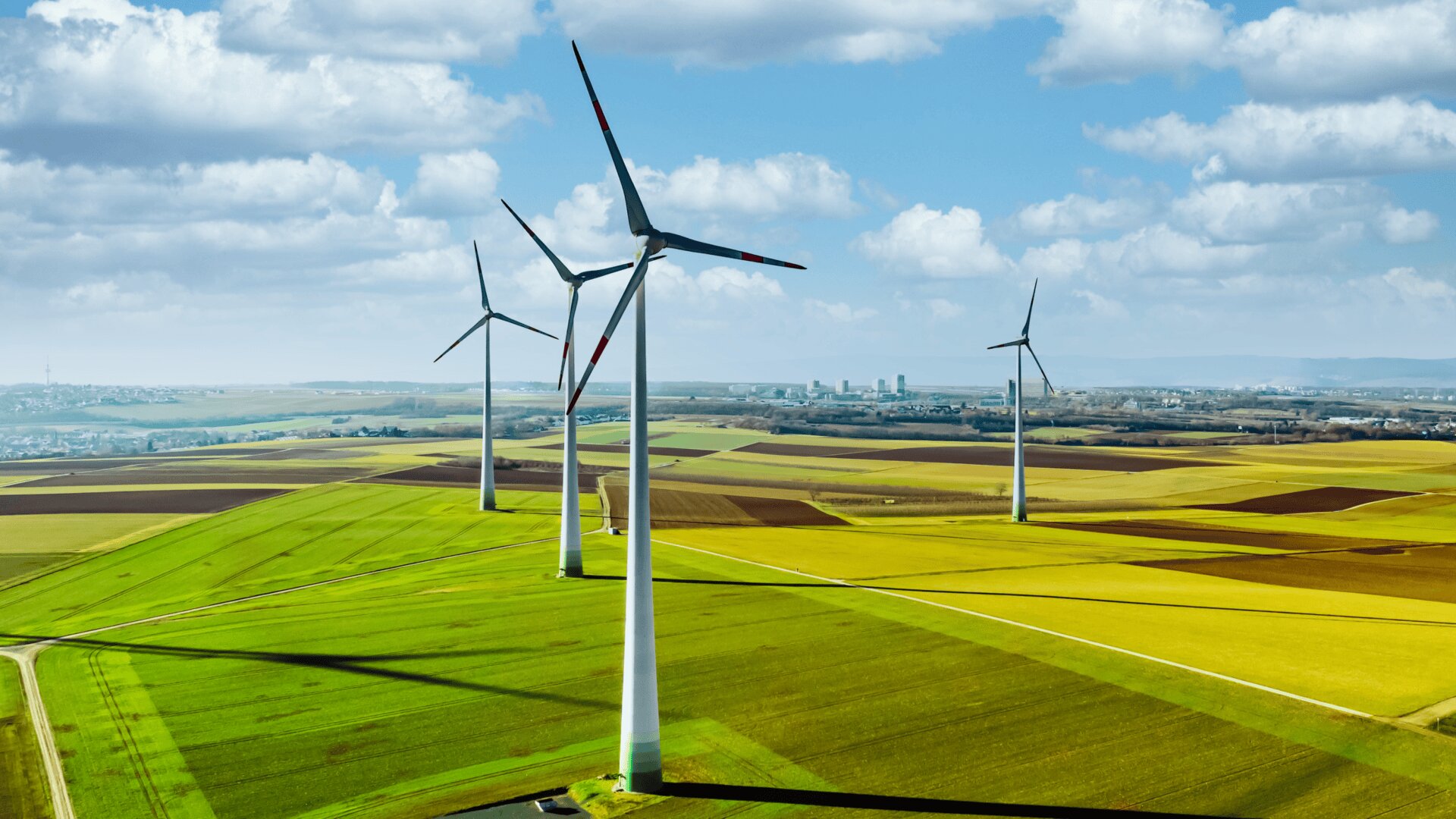 Drei große Windturbinen stehen in einem leuchtend grünen und gelben Flickenteppich aus Ackerland und drehen sich unter einem teilweise bewölkten blauen Himmel, an dessen Horizont eine entfernte Stadt zu sehen ist.