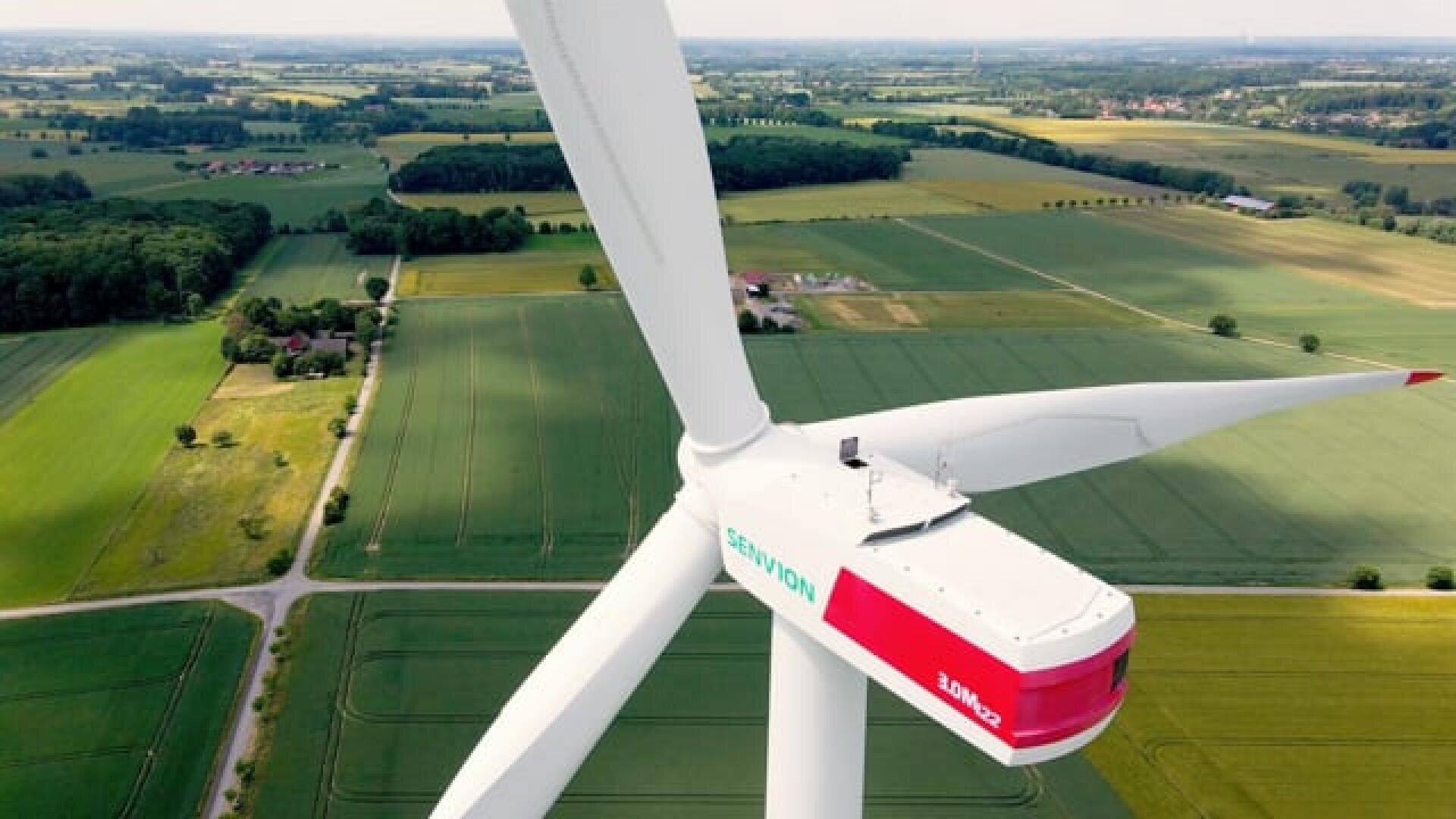 Nahaufnahme einer großen Windturbine mit weißen Flügeln, die bei klarem Himmel über grünes Ackerland, Felder und Straßen ragt. Das Wort Senvion ist auf der Turbinengondel sichtbar.