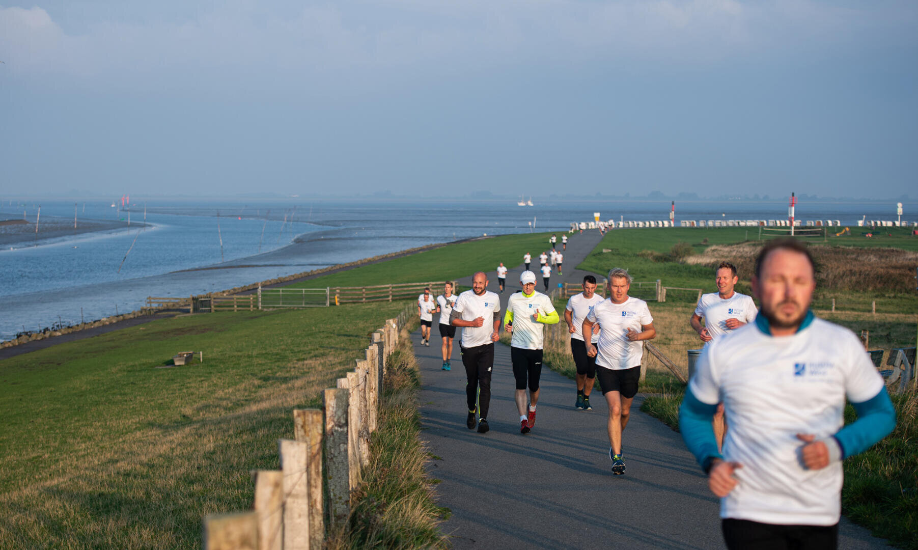 A group of people walk along a paved coastal path past green grass and water on a bright day with a clear blue sky. Everyone is wearing white shirts.