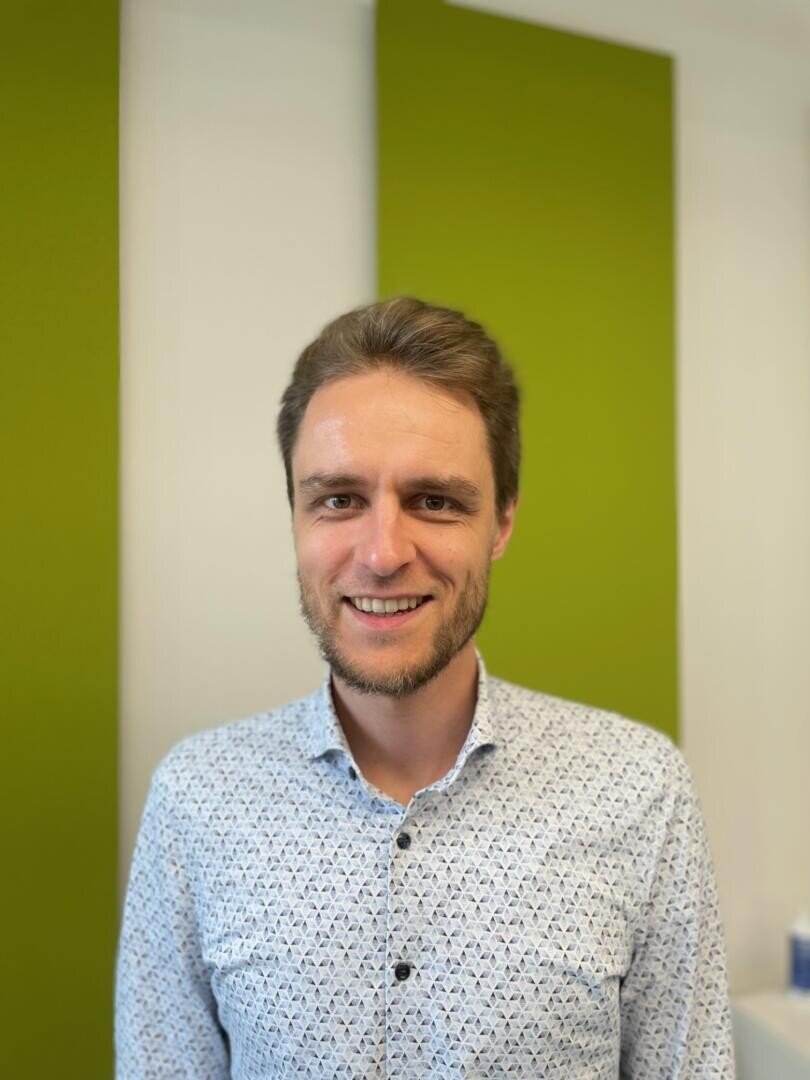 A man with short brown hair and a beard, wearing a light patterned shirt, stands smiling in front of a wall with two large vertical green panels. The background is bright and slightly blurred.