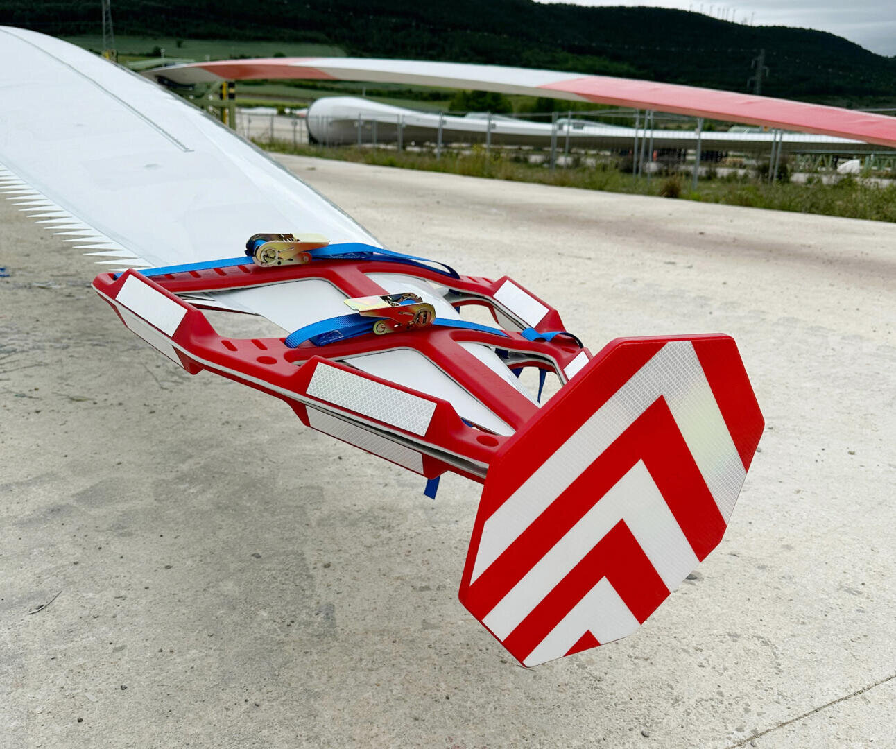 Close-up of the tip of a large wind turbine blade with a red and white chevron warning sign and securing straps, situated outdoors on a concrete surface with grassy hills in the background.