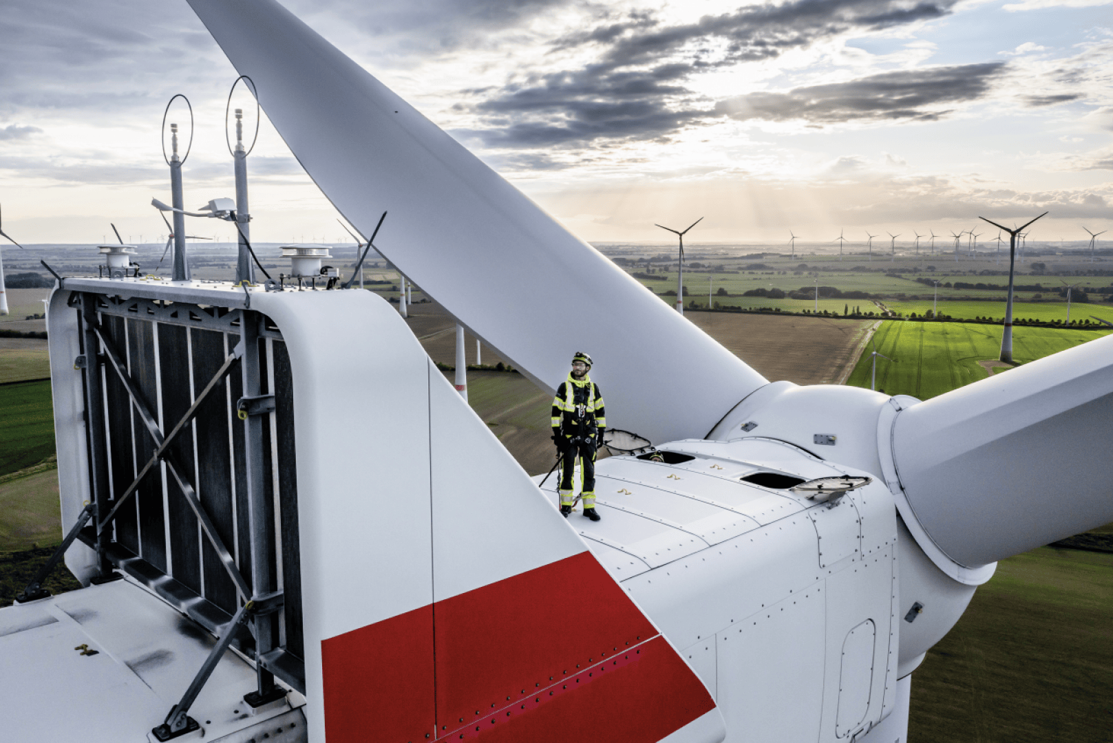 Ein Arbeiter in Schutzkleidung steht auf einer großen Windturbine und überblickt eine Landschaft mit grünen Feldern und mehreren Windturbinen unter einem teilweise bewölkten Himmel bei Sonnenuntergang.