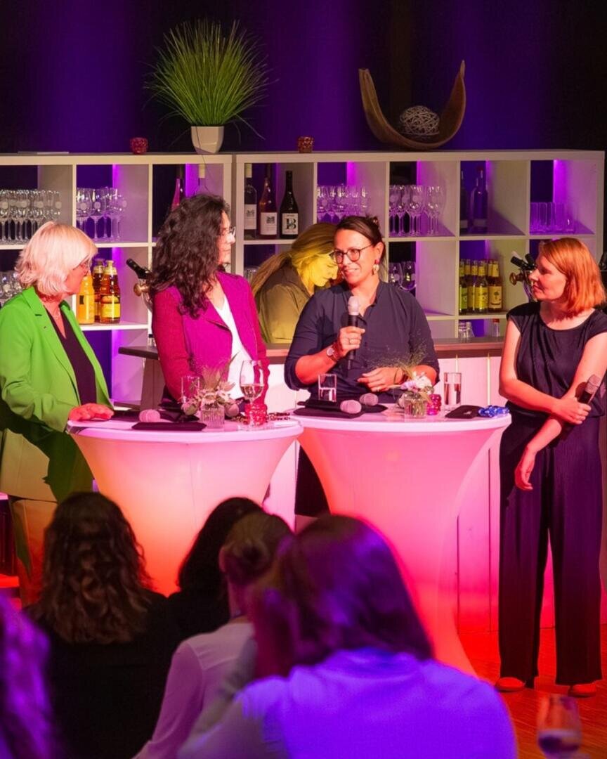 Vier Frauen stehen auf der Bühne an zwei runden, beleuchteten Cocktailtischen und sprechen während einer Podiumsdiskussion in Mikrofone. Im Hintergrund sind Regale mit Flaschen und Gläsern zu sehen. Im Vordergrund sind Zuschauer zu erkennen.