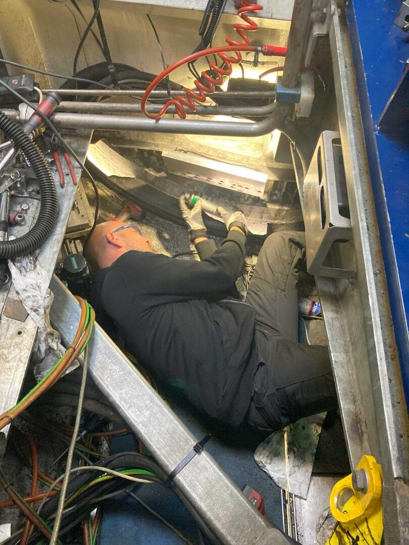 A person wearing gloves and dark clothing lies on their side in a cramped, space up in the wind turbine tower, working with tools and surrounded by cables, machinery parts, and bright overhead lighting. He is repairing the yaw ring