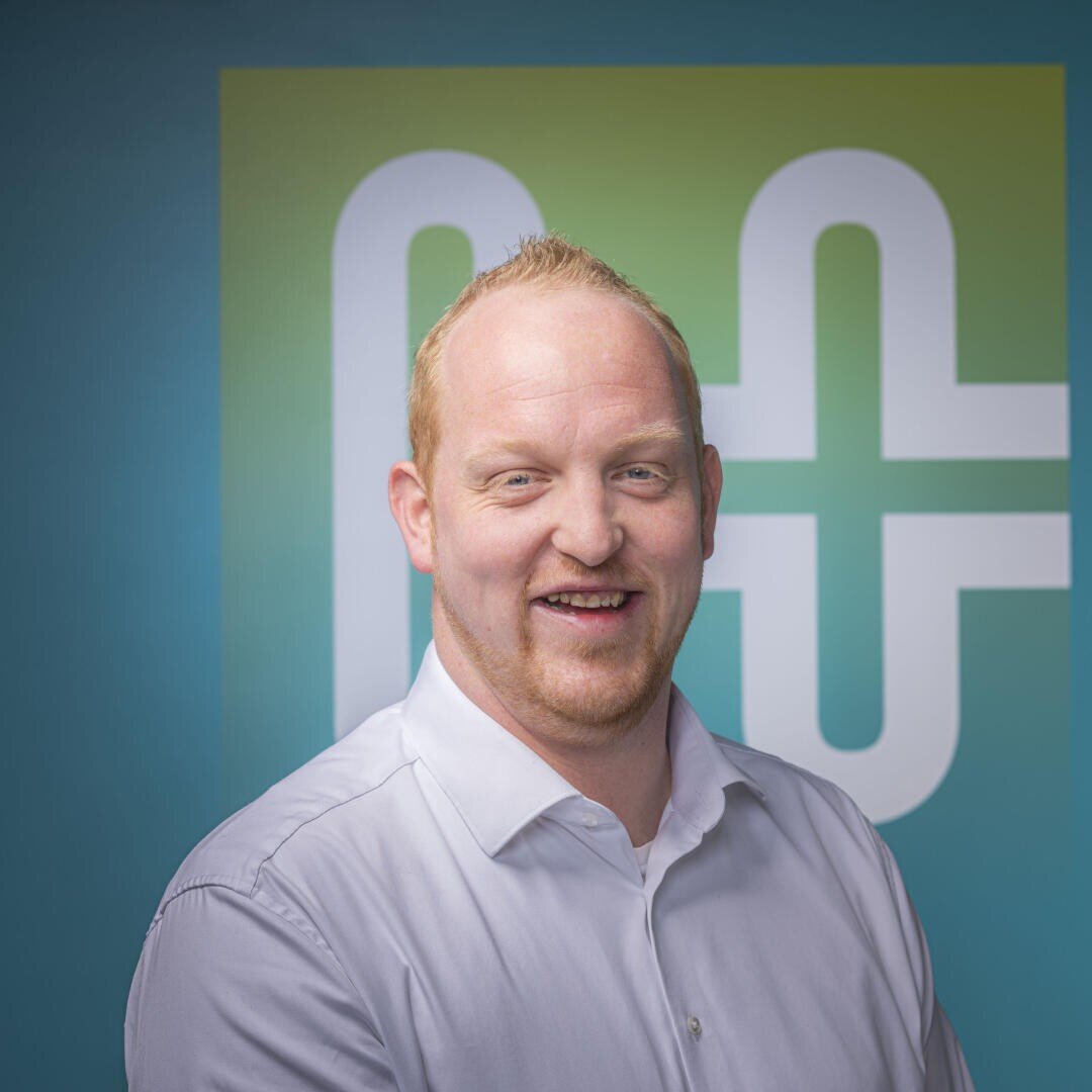 A smiling man with short blond hair wearing a white button-up shirt stands in front of a teal and green background with a white geometric logo.