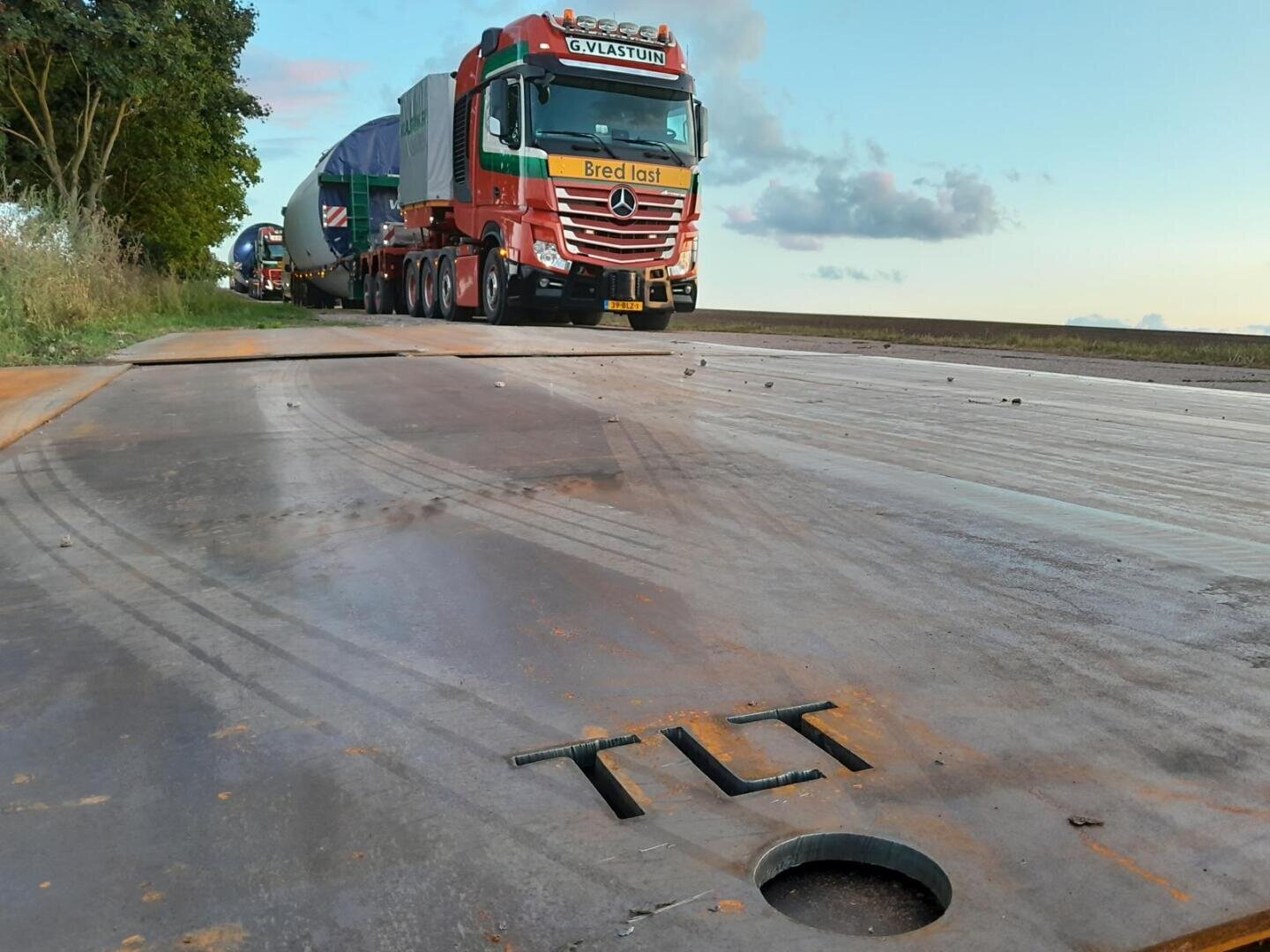 A large red truck transporting goods drives on a metal-plated road with TLT cut into the metal surface; another truck follows behind under a blue sky with scattered clouds.