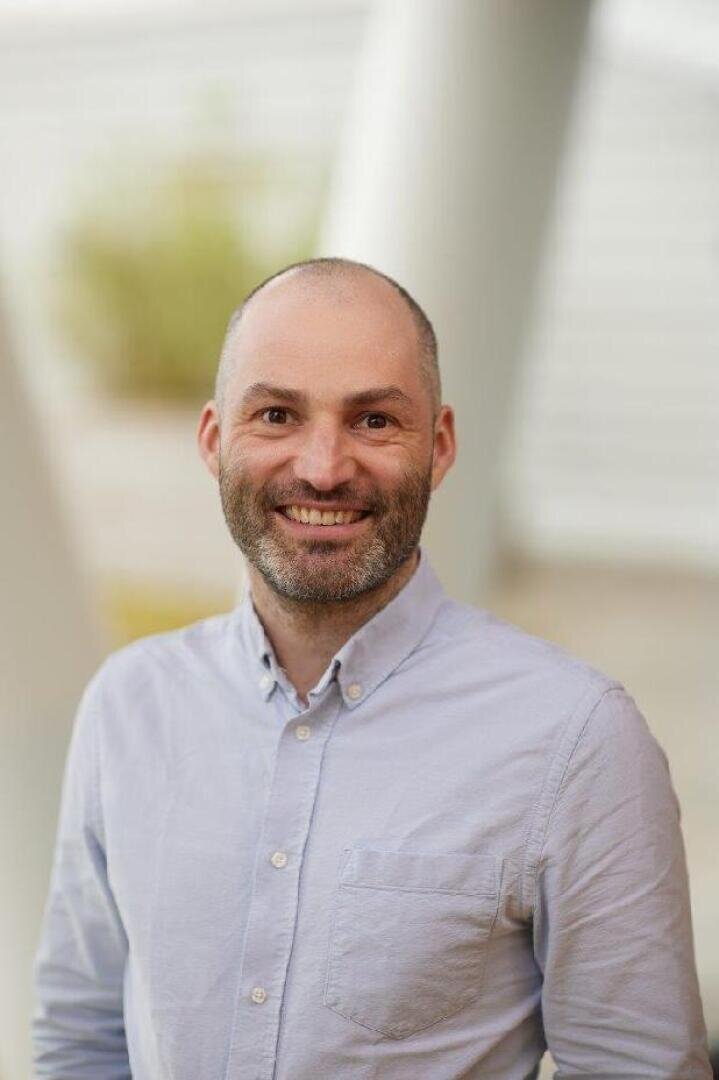 A smiling man with a beard and shaved head wears a light blue button-up shirt. He is standing outdoors in front of a blurred background with white structures and green plants.