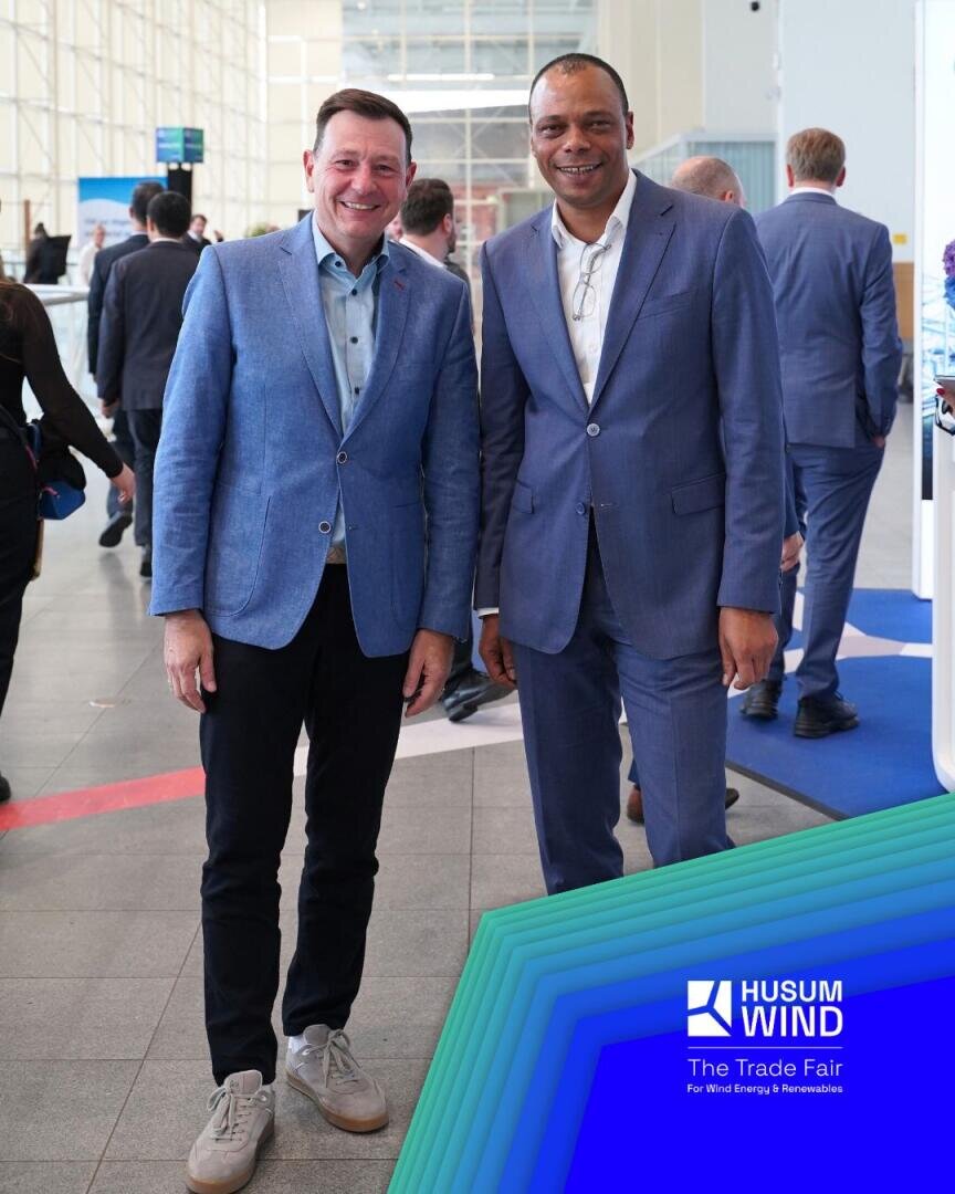 Zwei Männer in blauen Anzügen, die lächelnd nebeneinander auf einer Messe in einer Halle stehen. Das „Husum Wind“-Logo und das Veranstaltungsbranding sind in der rechten unteren Ecke zu sehen. Im Hintergrund sind Menschen zu sehen, die vorbeigehen.