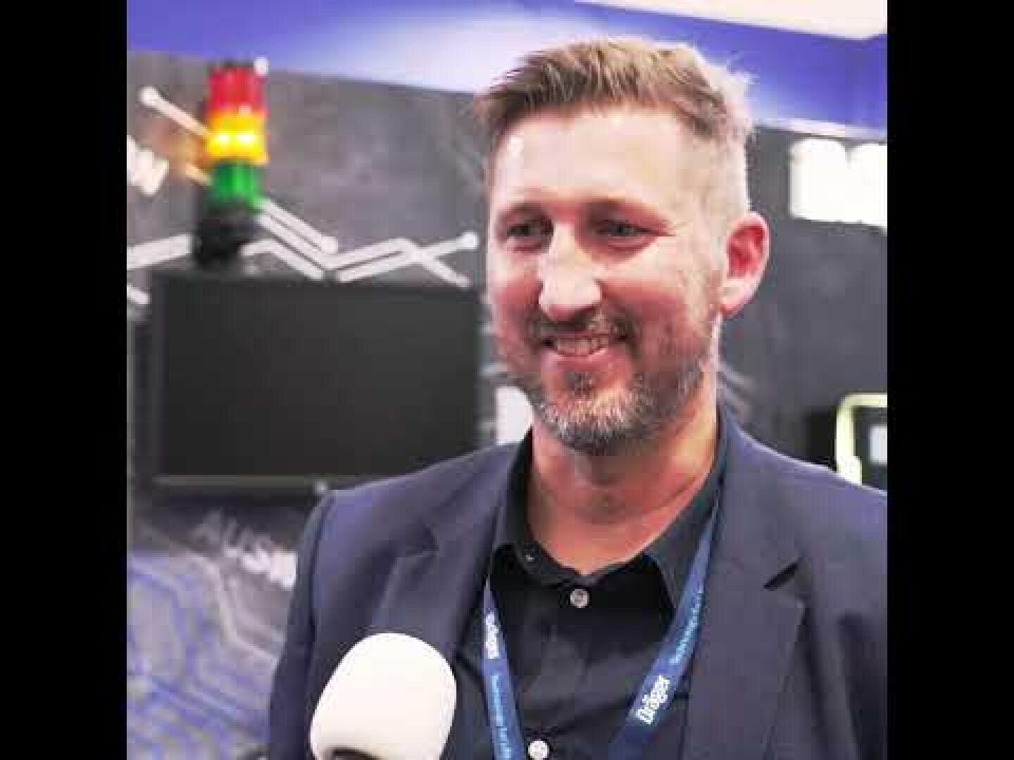 Ein Mann mit hellbraunem Haar und Bart, der einen marineblauen Blazer und ein Schlüsselband trägt, lächelt, während er mit einem Mikrofon vor sich interviewt wird. Im Hintergrund sind eine Tafel und eine Signallampe zu sehen.