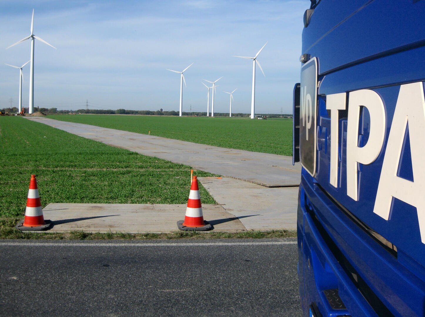 Ein blauer Lastwagen parkt am Straßenrand in der Nähe von orangefarbenen Verkehrsleitkegeln, während im Hintergrund mehrere Windkraftanlagen auf einer grünen Wiese unter einem klaren Himmel stehen.
