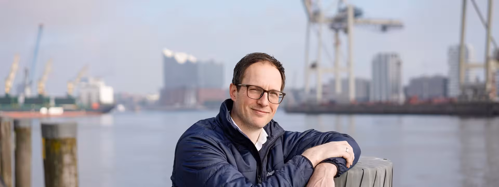 A man with glasses and a navy blue jacket leans against a wooden post by the water. Cranes, industrial buildings and the city skyline can be seen in the background.