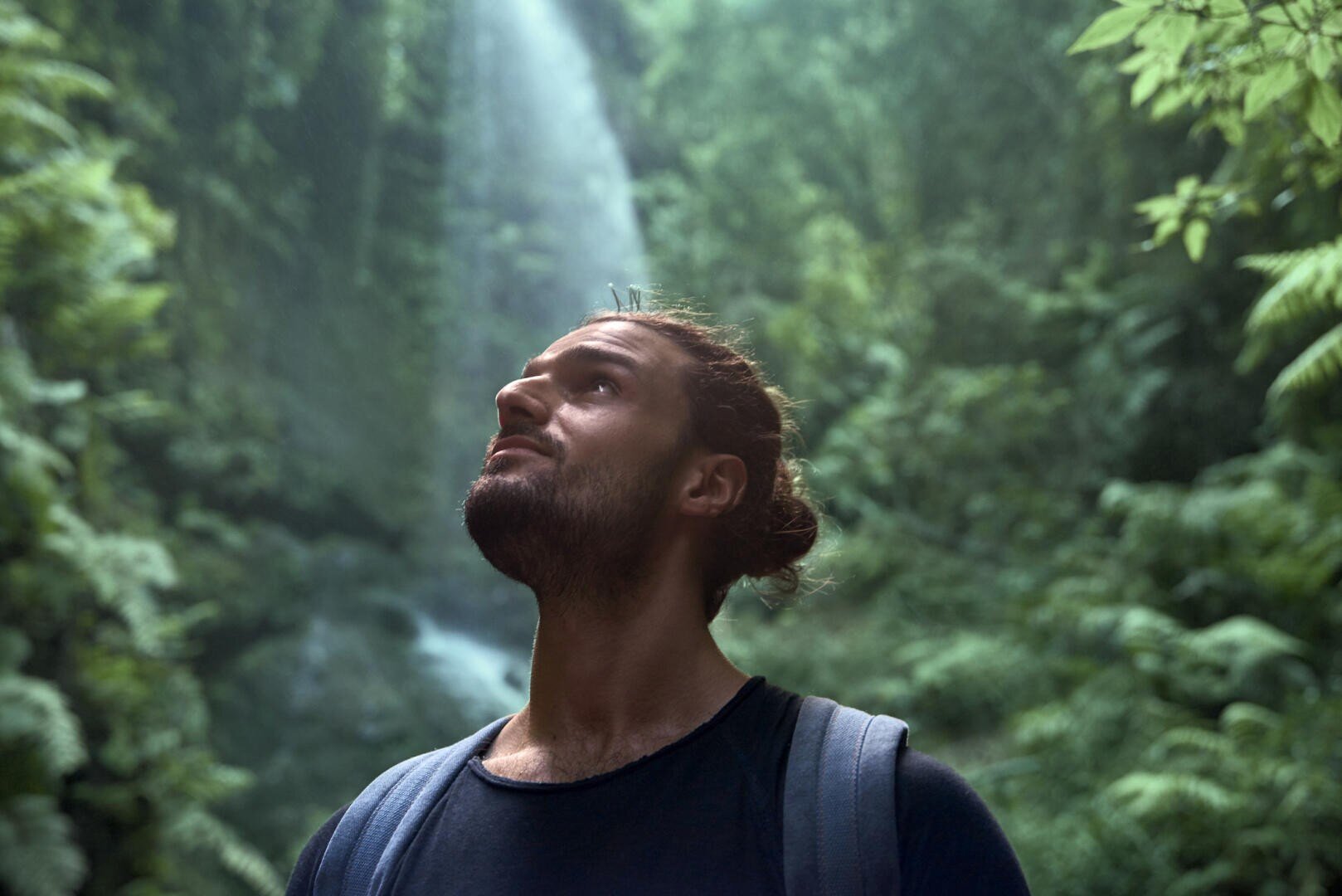 A man with a beard and his hair tied back looks up as he stands in a lush, green forest with a waterfall in the background. The sunlight falls through the trees above him.