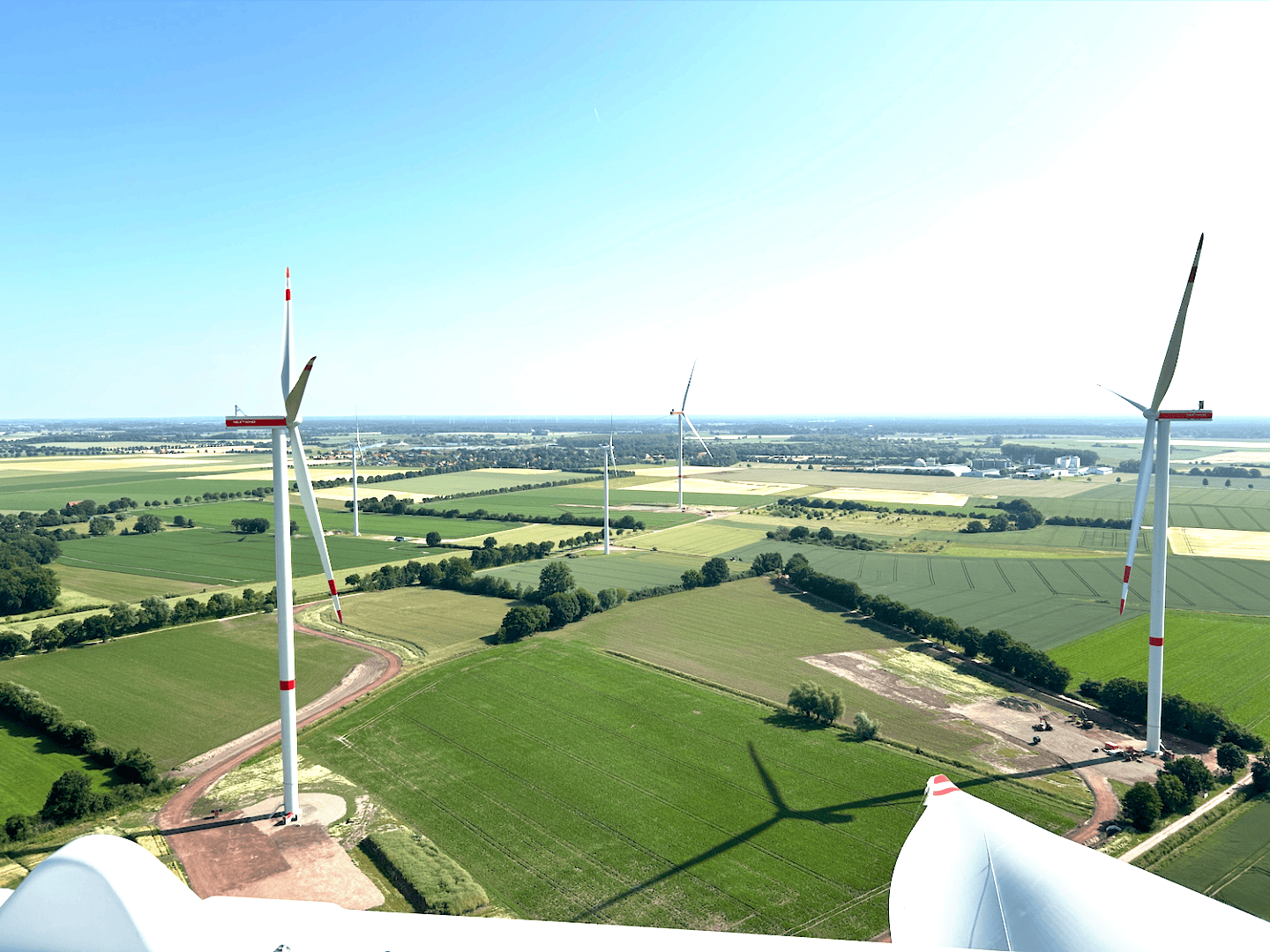 Luftaufnahme mehrerer Windturbinen, die sich über grüne Felder und Ackerland unter einem klaren blauen Himmel ausbreiten, wobei in der Ferne Gebäude und Bäume am Horizont zu sehen sind.