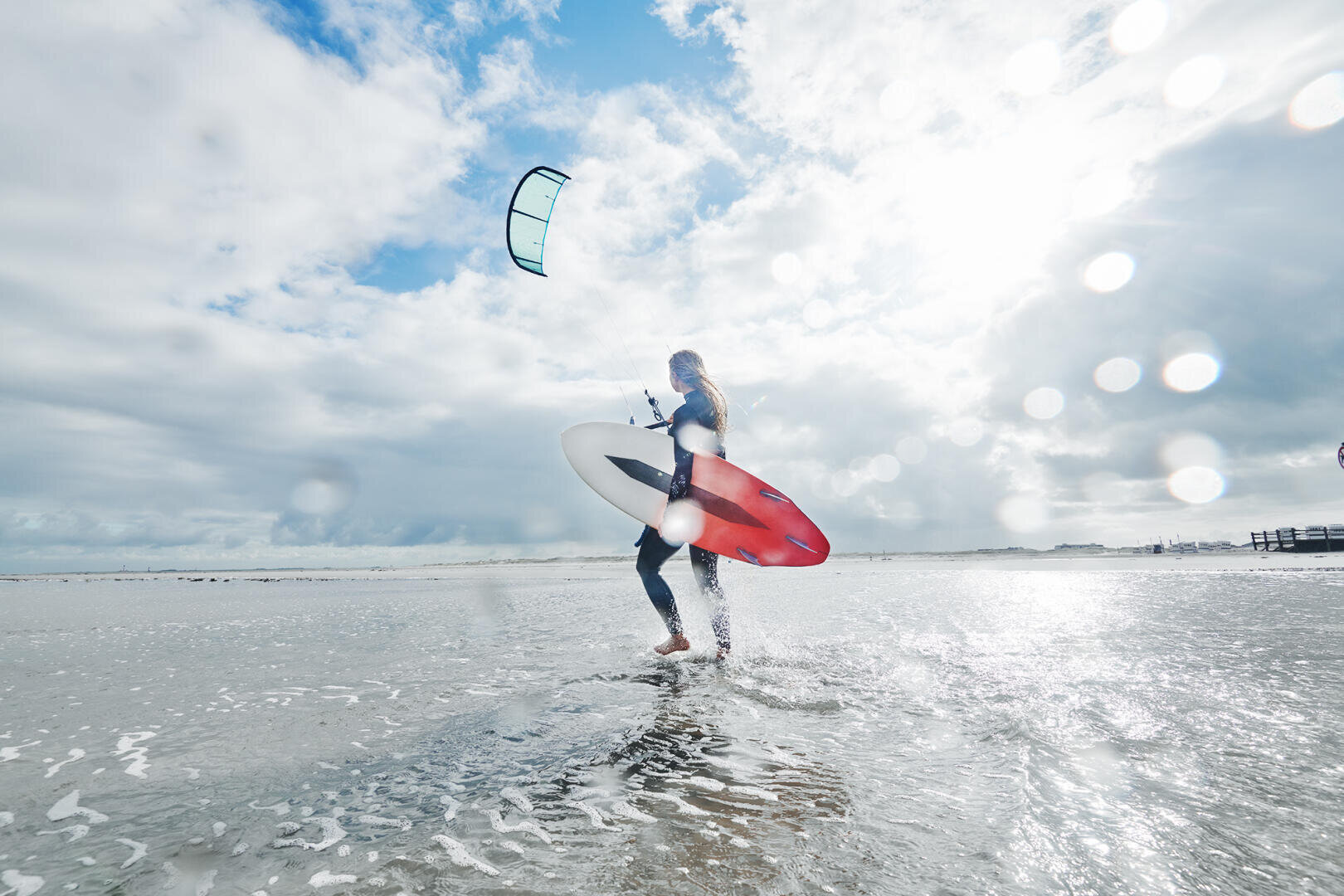 Eine Person steht an einem nassen Strand und hält ein rot-weißes Surfbrett, über dem ein Drachen schwebt. Die Szene ist hell mit vereinzelten Wolken und Sonnenlicht, das sich auf dem Wasser spiegelt.