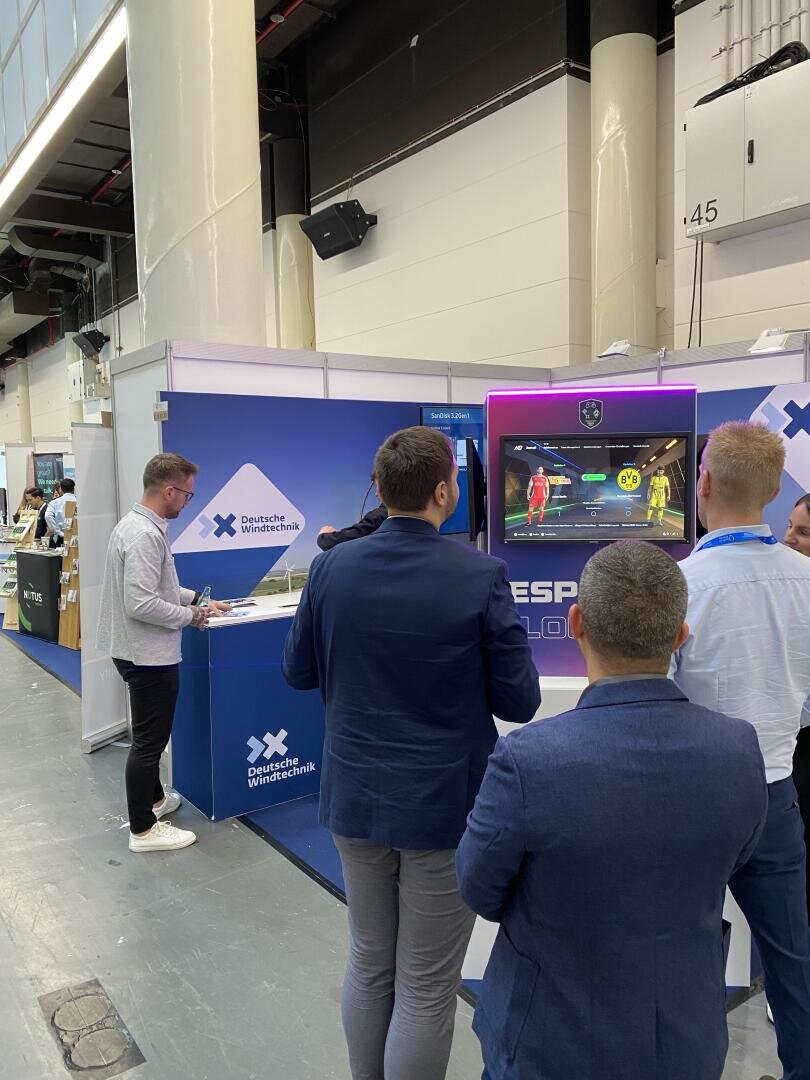 Mehrere Männer in Businesskleidung schauen oder spielen ein Fußballvideospiel an einem Stand mit der Aufschrift „Deutsche Wirtschaft“ in einem großen Veranstaltungsraum. Ein Mann steht mit einem Laptop hinter einem blauen Tresen.