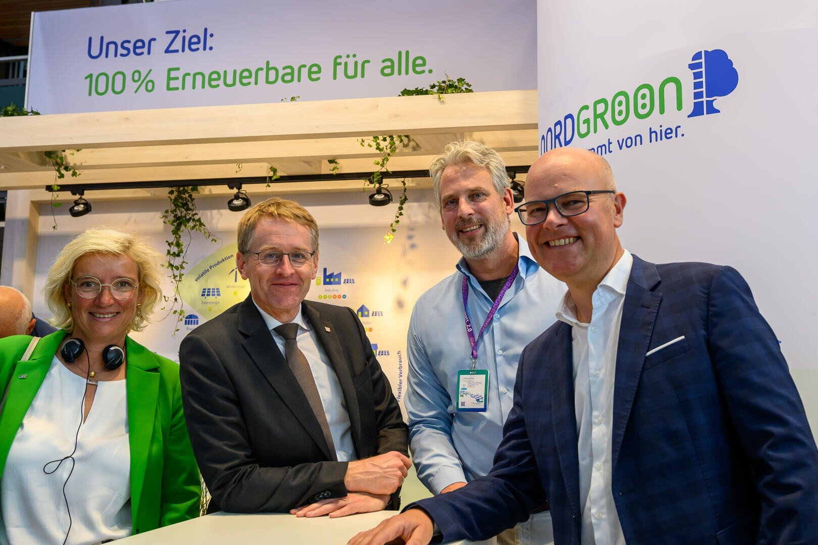 Vier lächelnde Menschen, drei Männer und eine Frau, stehen zusammen an einem Veranstaltungsstand. Hinter ihnen steht ein deutsches Schild mit der Aufschrift "Unser Ziel: 100% Erneuerbare für alle".