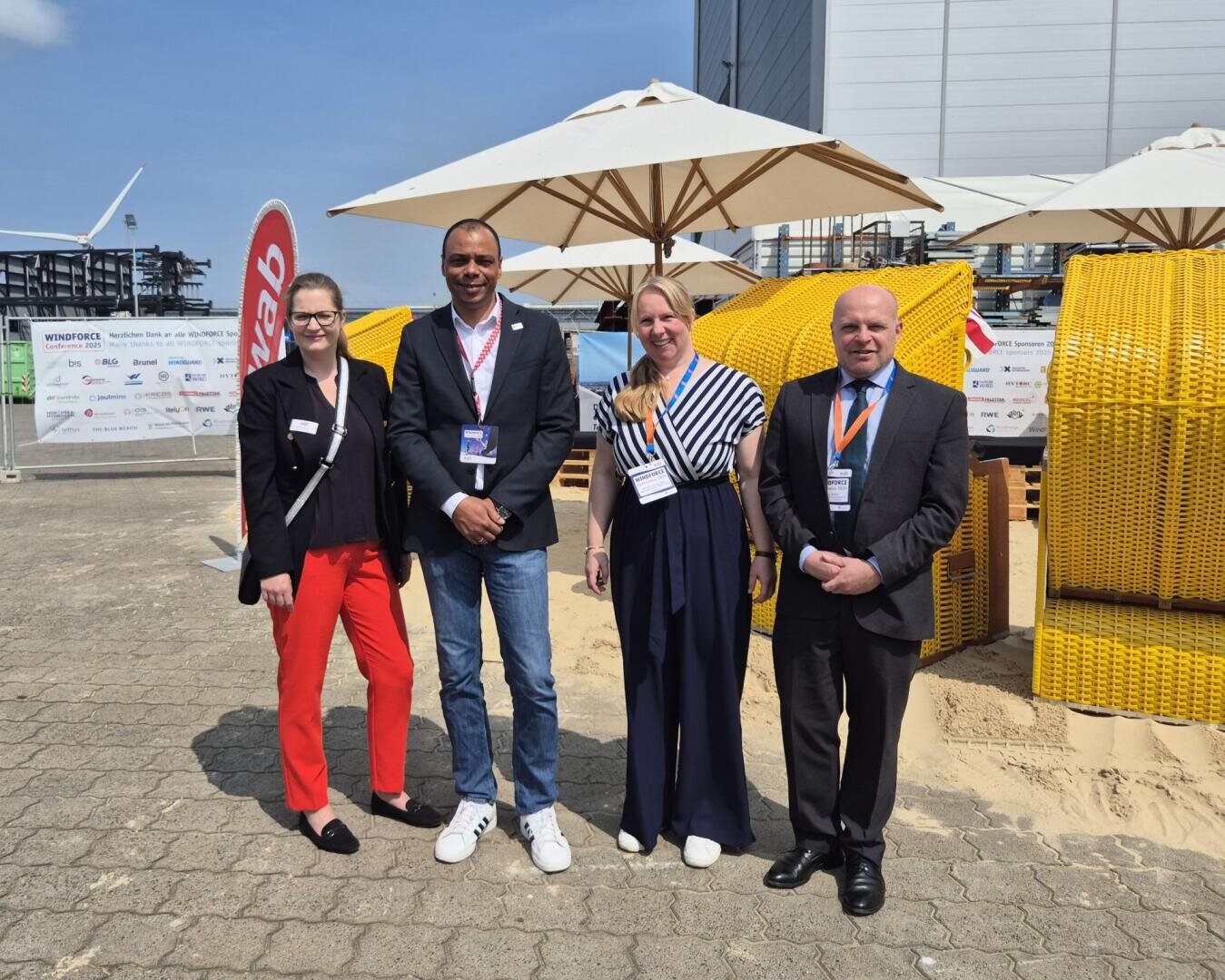 Vier Personen stehen lächelnd in Business-Kleidung vor gelben Strandkörben und großen Sonnenschirmen, mit einem Gebäude, Veranstaltungsschildern und blauem Himmel im Hintergrund.