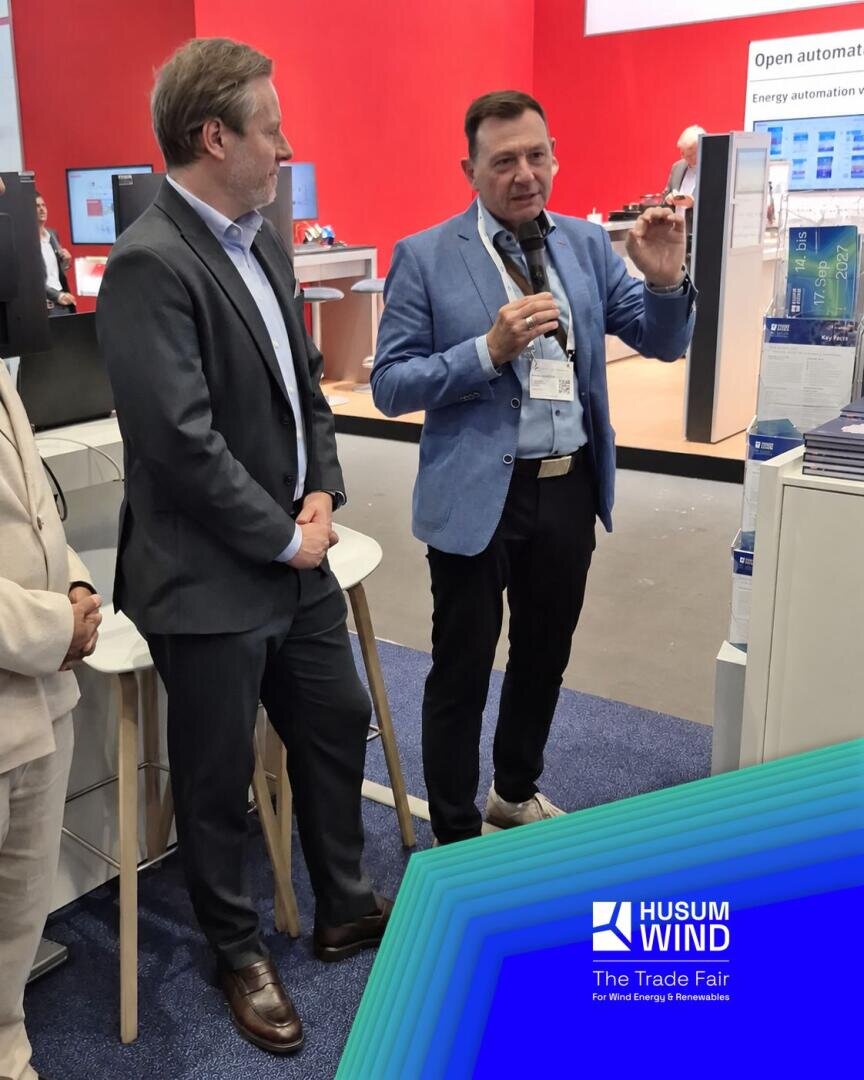 Zwei Männer in Geschäftskleidung stehen auf einem blauen Teppich an einem Messestand. Einer hält ein Mikrofon in der Hand und gestikuliert beim Sprechen. In der rechten unteren Ecke ist das Logo der Husum Wind-Messe zu sehen.