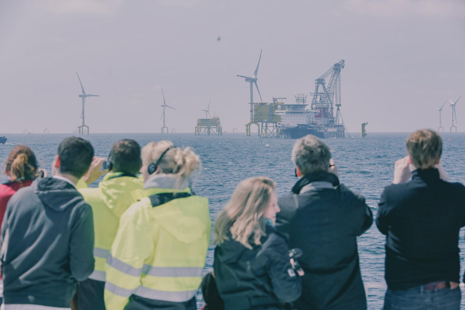 A group of people in light-colored jackets stand at the water's edge, observing and photographing offshore wind turbines and industrial platforms in the distance under a cloudy sky.