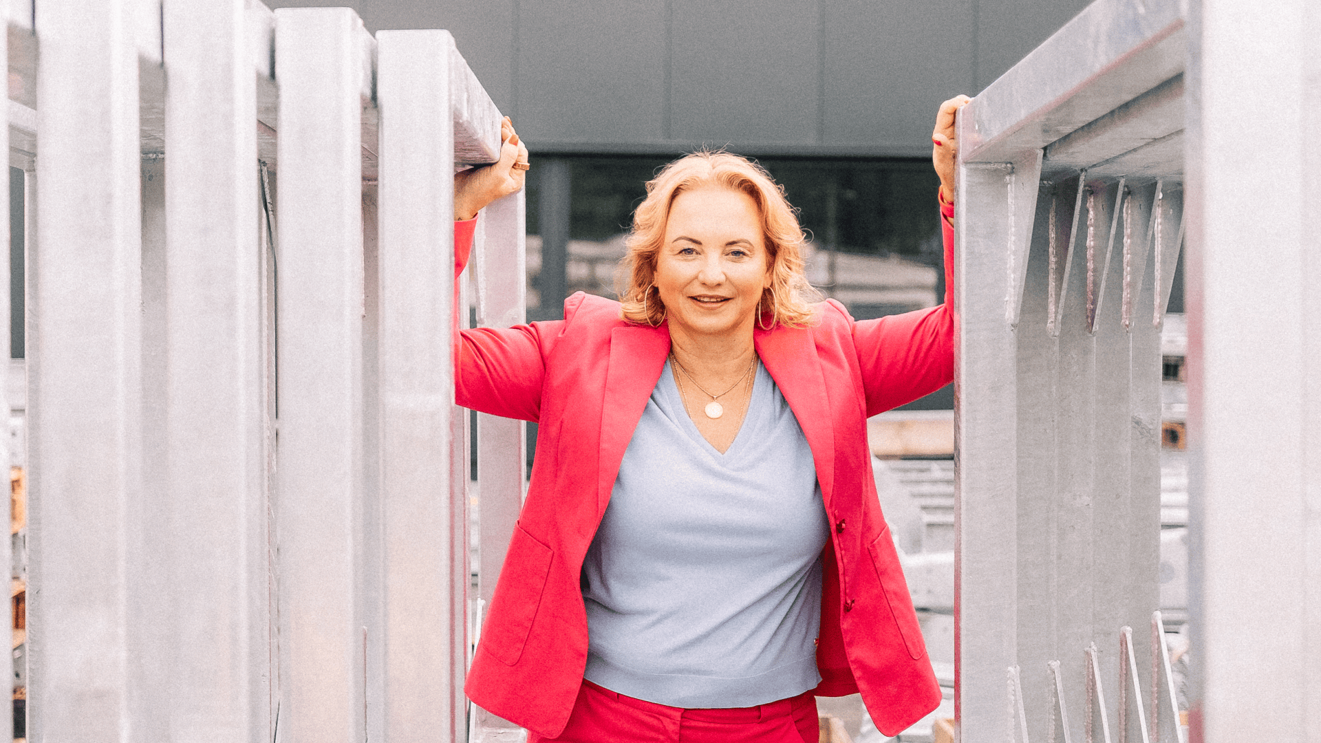 A woman in a bright pink suit and light blue top stands confidently outdoors, holding on to two large metal supports on either side, while a modern building can be seen in the background.