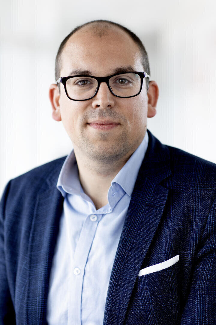 A man with glasses, a blue blazer, a light blue shirt and a white handkerchief sits opposite the camera in front of a bright, blurred background.