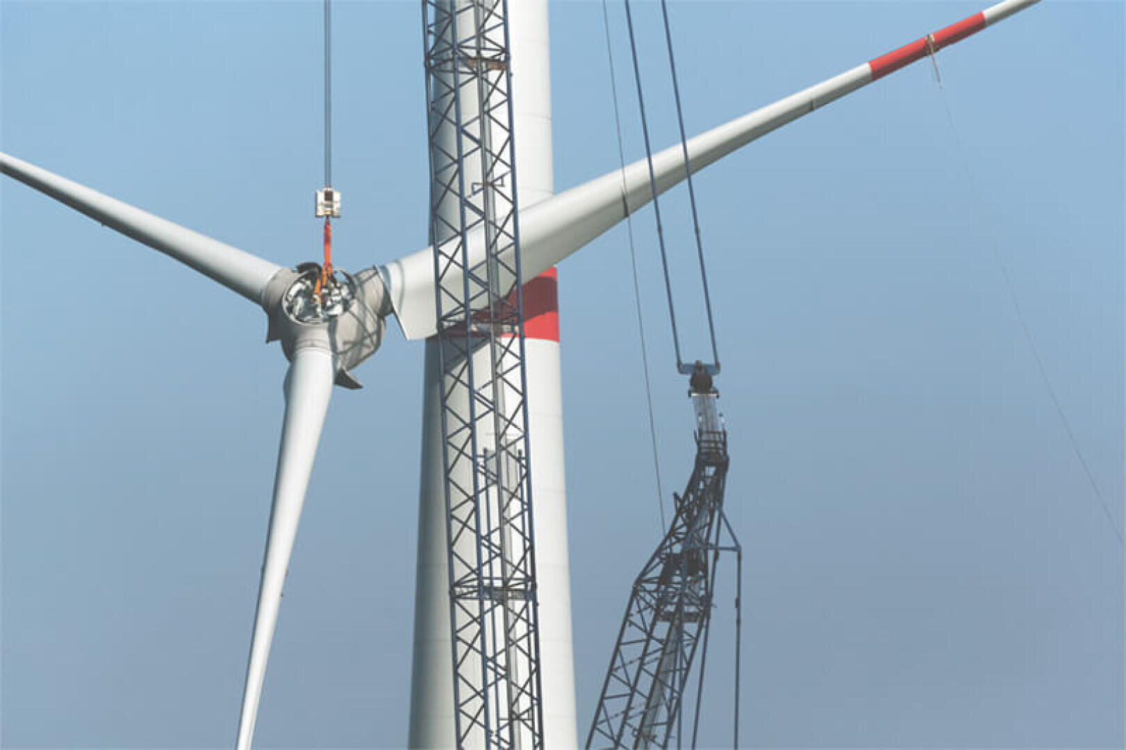 Ein großer Kran montiert bei strahlend blauem Himmel die Rotorblätter auf dem Turm einer Windkraftanlage und demonstriert damit einen Teil des Bauprozesses von Windkraftanlagen.