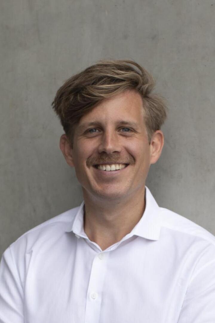 A man with light brown hair and a mustache, wearing a white button-up shirt, stands smiling in front of a plain gray background.