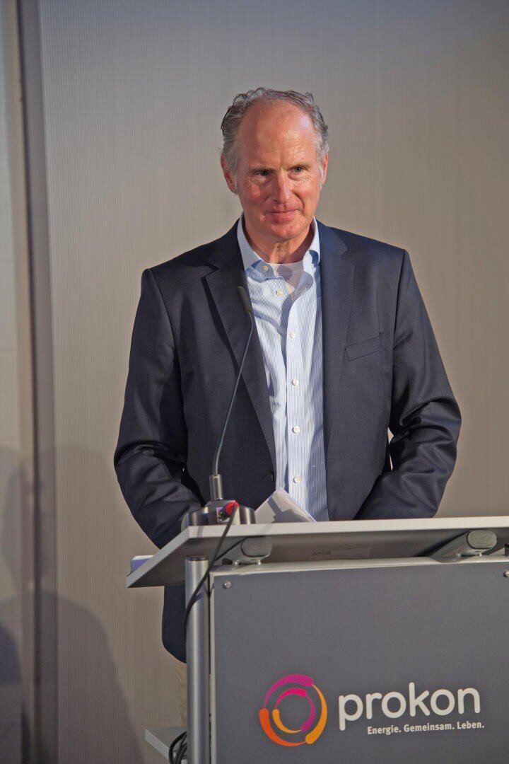 A man in a suit stands at a podium with the prokon logo, holding papers and speaking in front of a plain background.