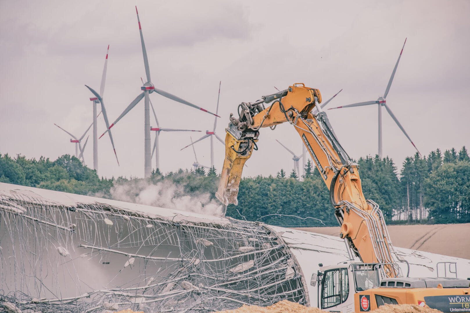 Ein gelber Bagger zerstört das Rotorblatt einer Windkraftanlage. Im Hintergrund sind unter einem bewölkten Himmel Windräder und Bäume zu sehen.
