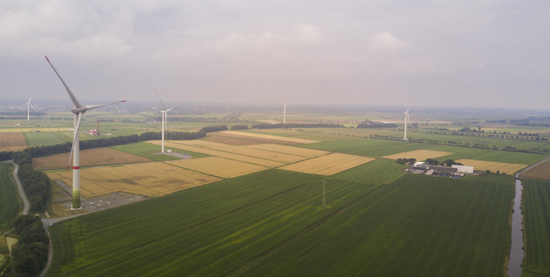 Luftaufnahme von grünem und gelbem Ackerland mit mehreren großen Windturbinen, die unter einem bewölkten Himmel über die Felder verteilt sind. In der Nähe des rechten Bildrandes sind einige Gebäude zu sehen.