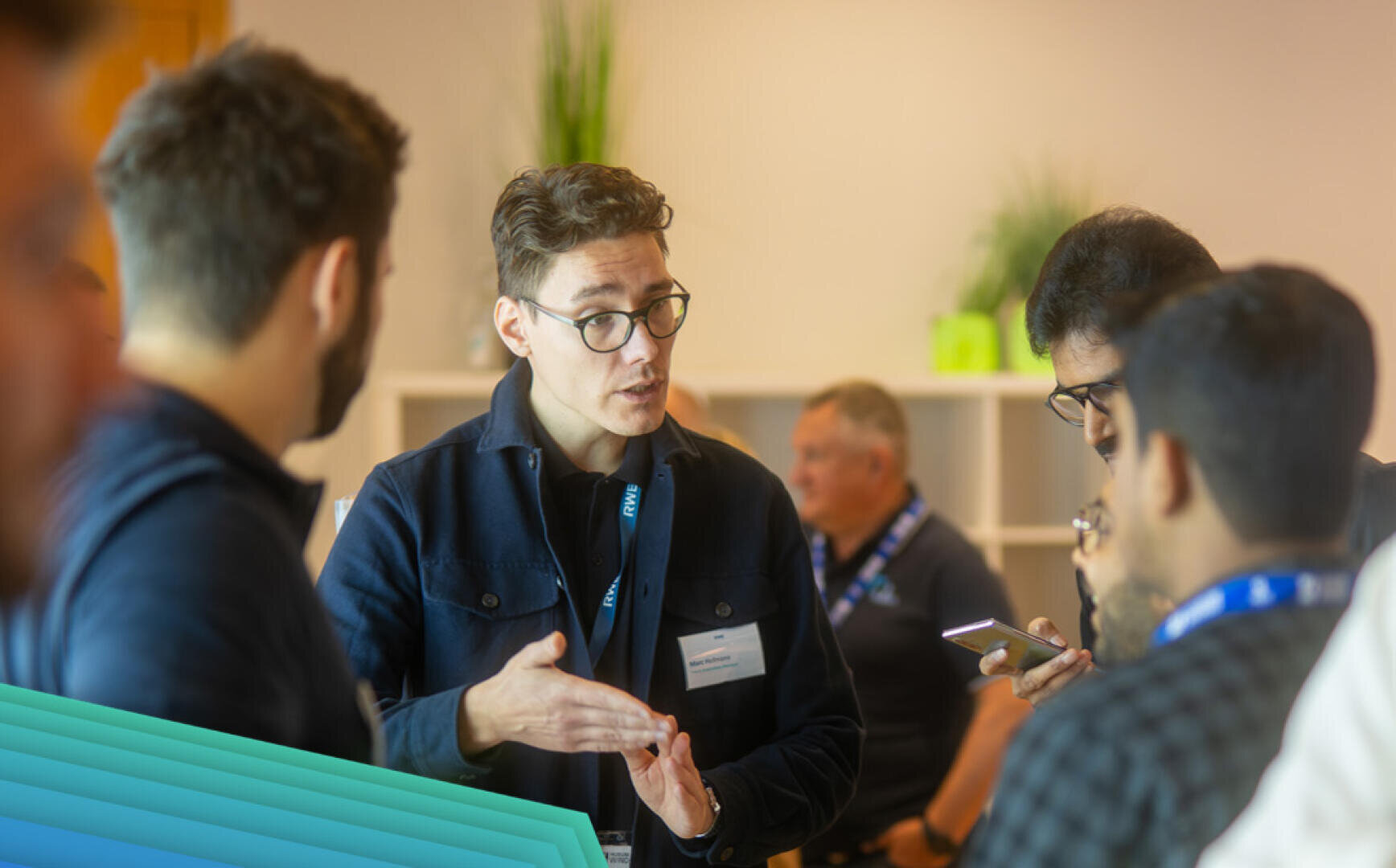 A group of people are talking at an event in a building. One person, wearing glasses, gestures while talking, while the others listen and look at their phones. Shelves and plants can be seen in the background.