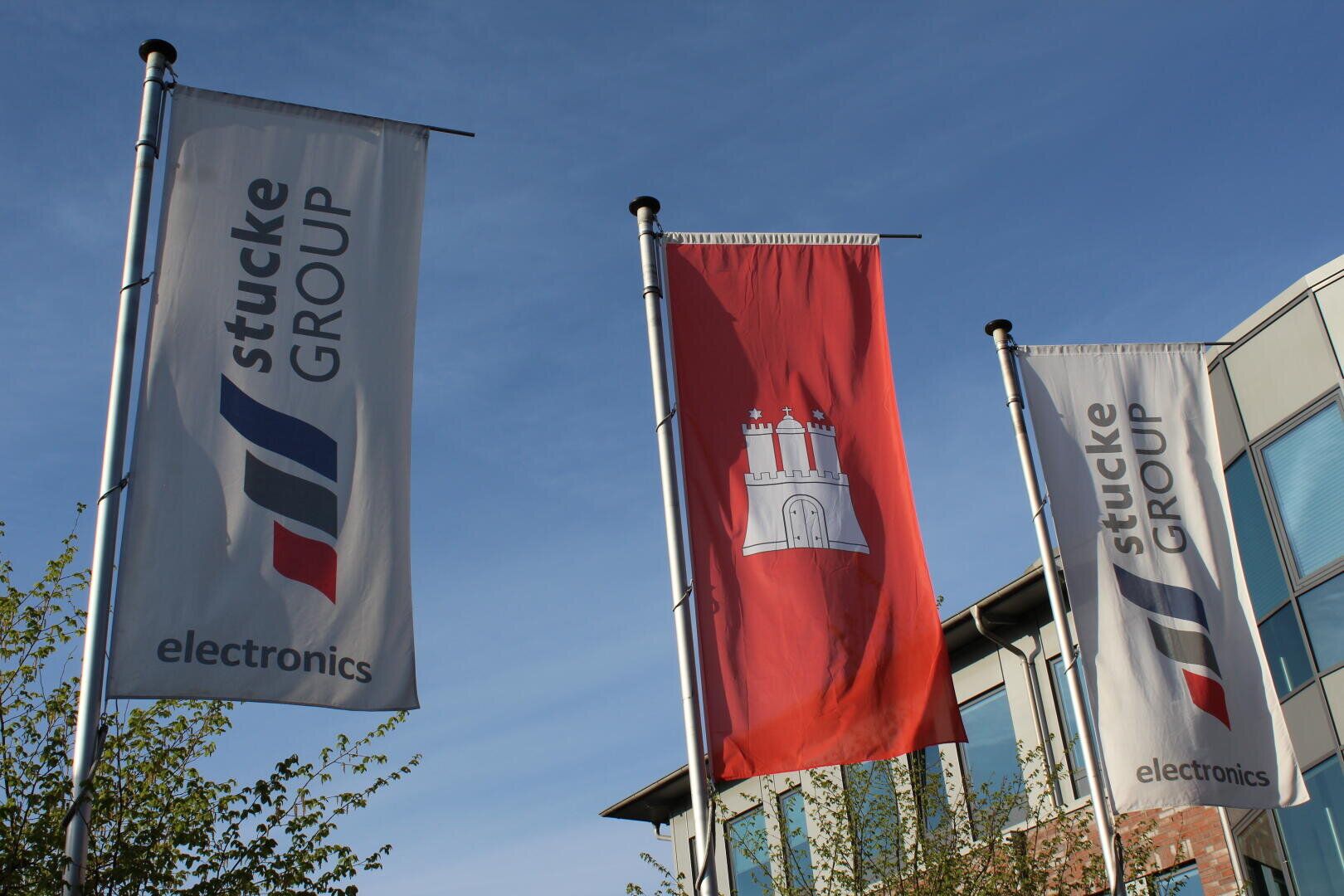 Drei Fahnen an Masten: zwei mit der Aufschrift "Stucke GROUP electronics" in Blau und Grau, und die mittlere Fahne ist rot mit einem weißen Schlosswappen. Im Hintergrund sind ein Gebäude und ein blauer Himmel zu sehen.