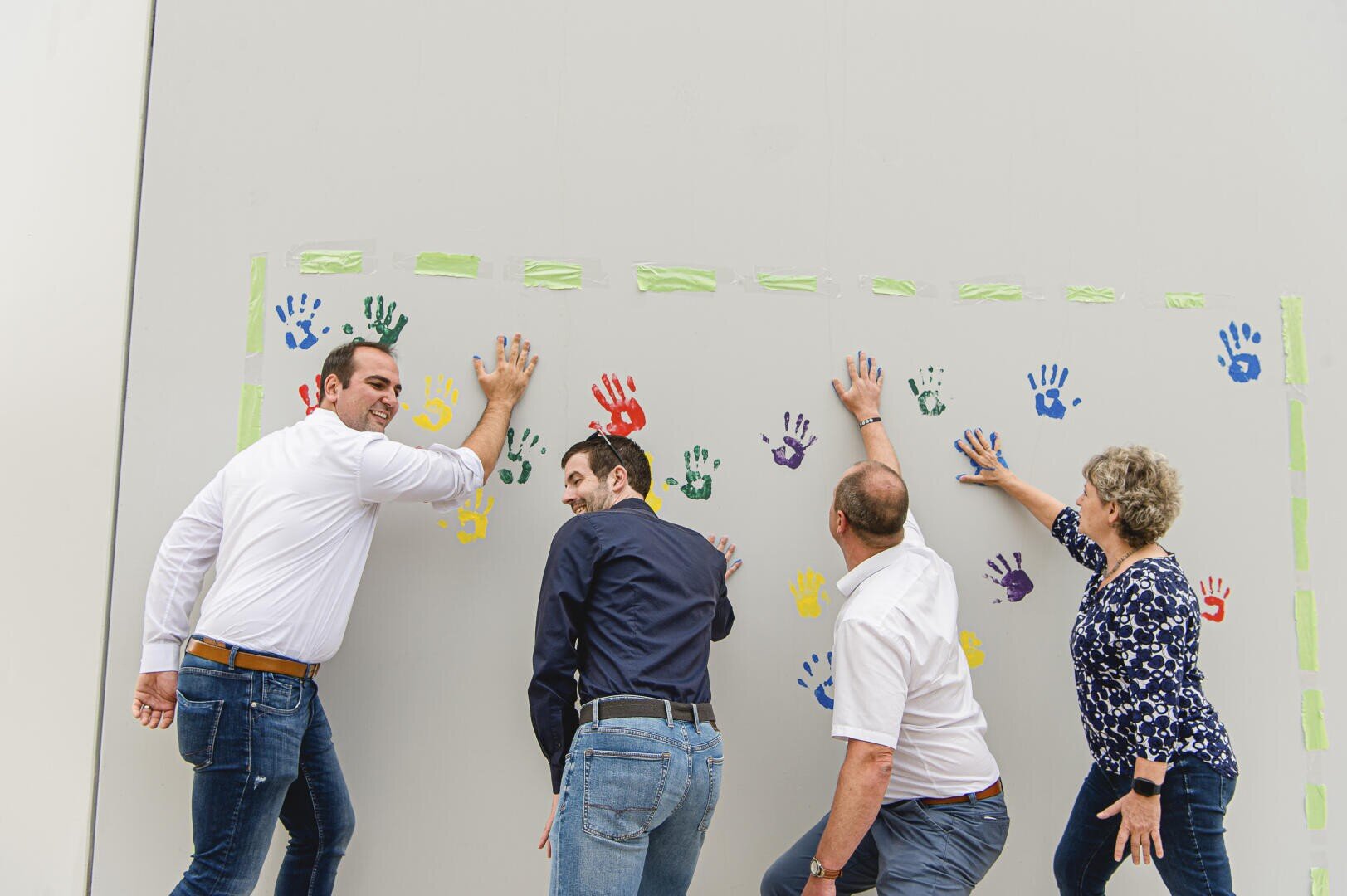 Vier Erwachsene drücken ihre bemalten Hände auf eine weiße Wand und fügen bunte Handabdrücke in einer Reihe hinzu. Sie stehen lächelnd und in Freizeitkleidung da und nehmen an einer Gruppenaktivität teil.