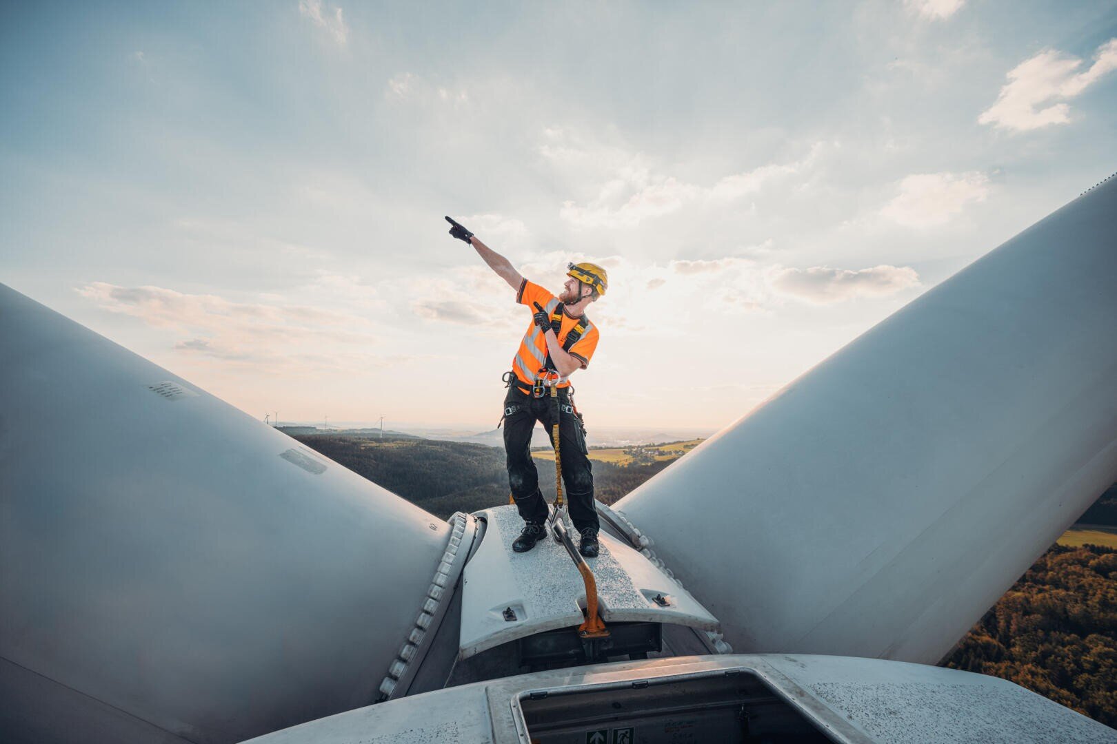 Ein Arbeiter in Schutzkleidung steht auf einer Windkraftanlage und zeigt mit einem Arm nach oben. Im Hintergrund sind die großen weißen Rotorblätter der Anlage und eine malerische Landschaft unter einem teilweise bewölkten Himmel zu sehen.