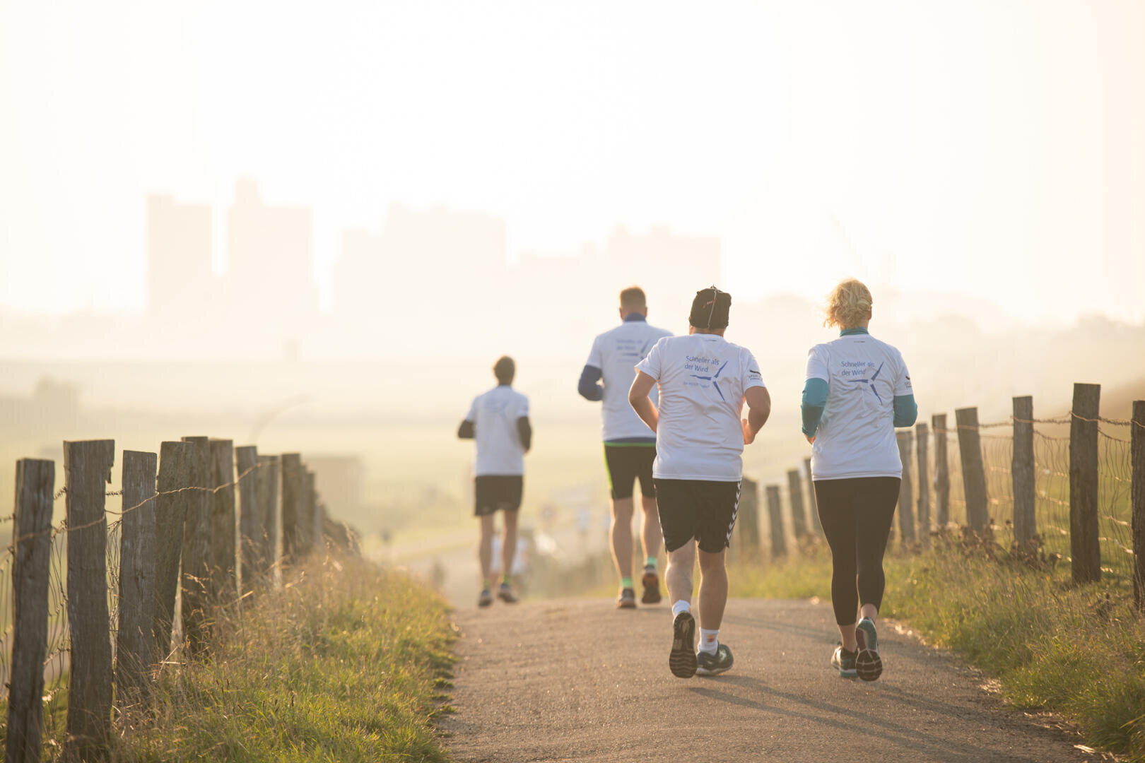 Four people in matching white shirts walk together along a country path lined with wooden fences toward the skyline of a distant city in the hazy sunlight.