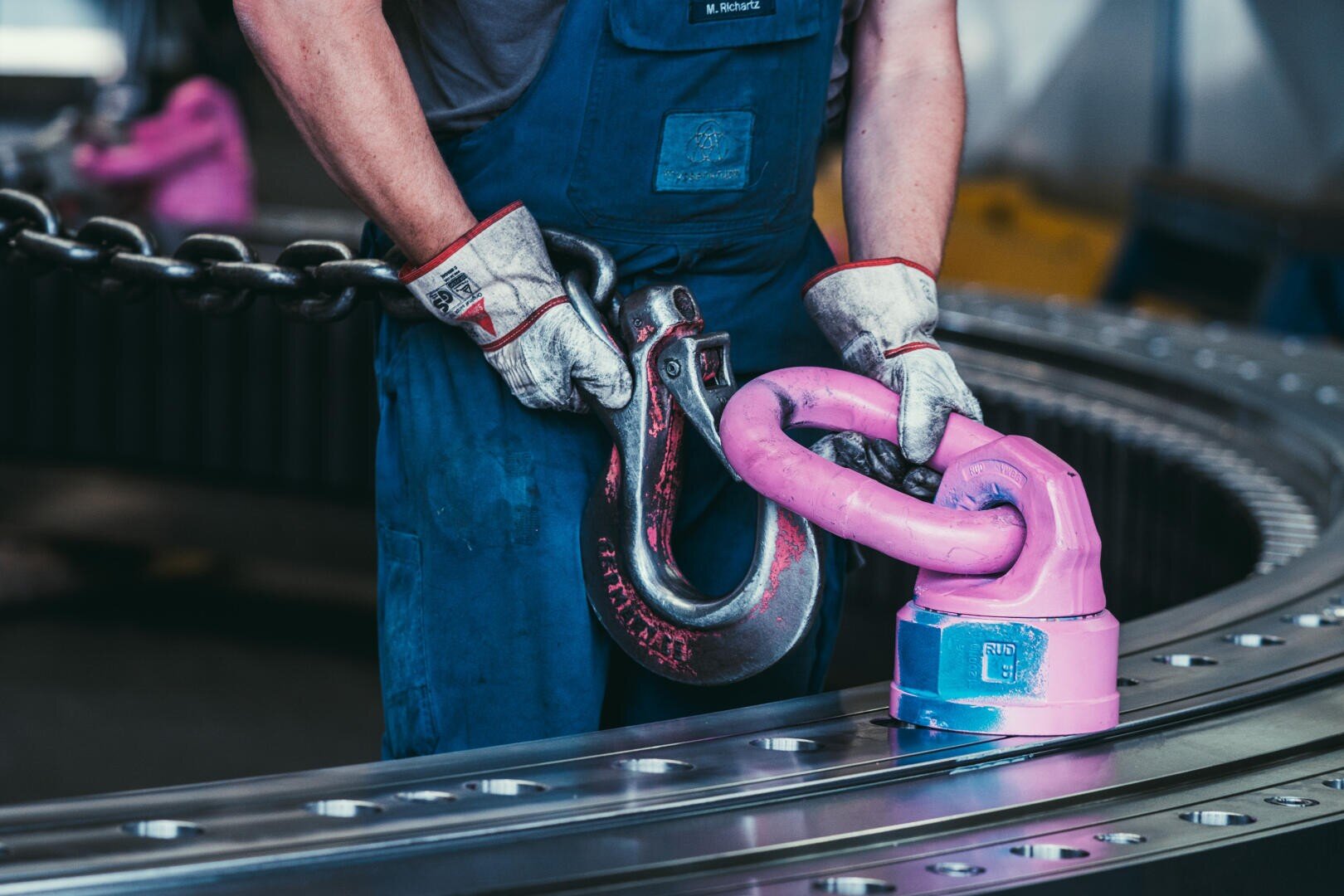 Ein Arbeiter mit Handschuhen und blauem Overall befestigt einen großen rosafarbenen Hebehaken an einer Metallkette in einer Industrieumgebung, mit einer gebogenen Metallstruktur im Hintergrund.