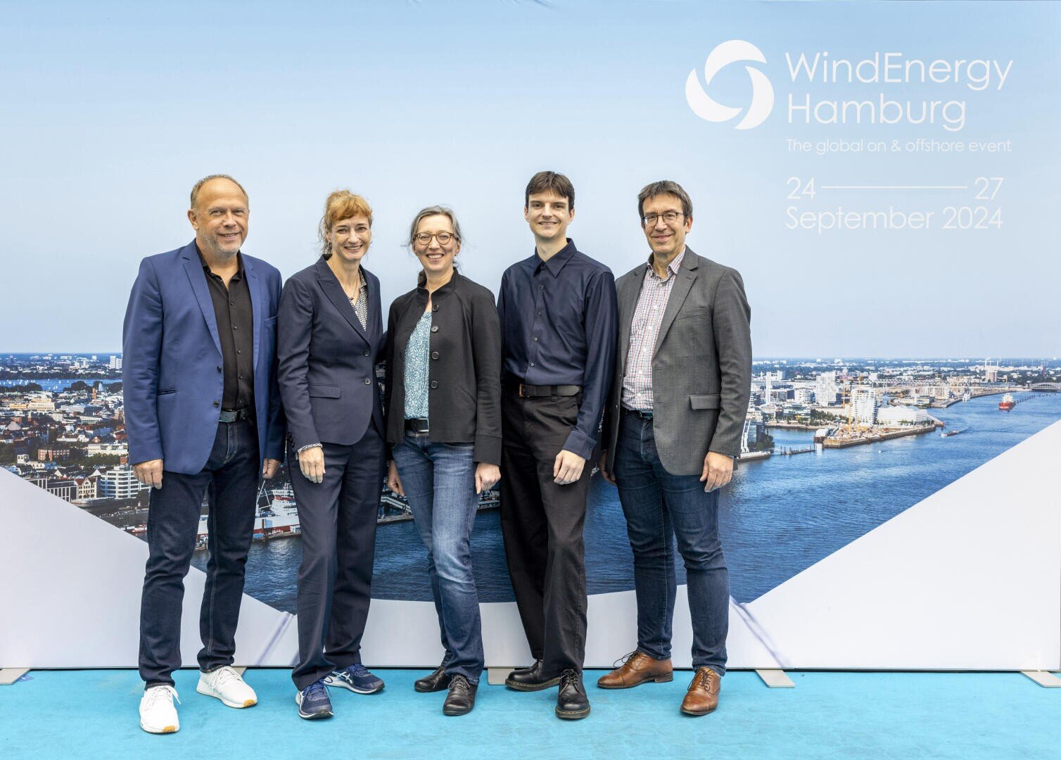 Fünf Personen stehen lächelnd vor einem Hintergrund, der eine Stadtlandschaft und Wasser zeigt, mit dem Text WindEnergy Hamburg und dem Veranstaltungsdatum 24-27 September 2024 in der oberen rechten Ecke.